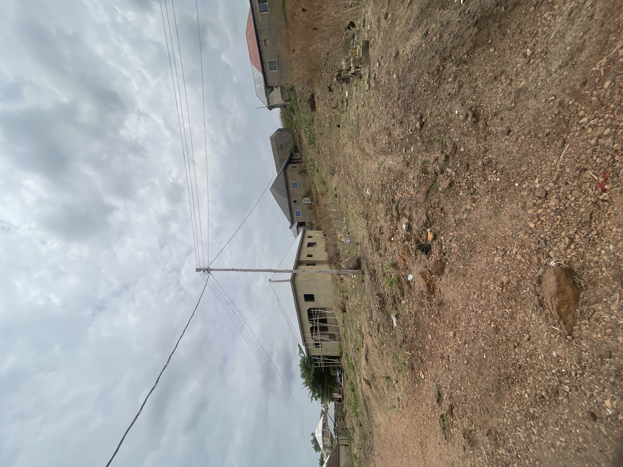 The image depicts a dirt road in Lokoja, Nigeria, ascending a slight incline. The ground is predominantly brown dirt with small rocks and sparse vegetation. Several unfinished or partially constructed buildings are visible along the hillside, indicating an area under development. Overhead power lines extend across the frame, supported by a wooden pole. The sky is overcast with grey clouds. No people are visible in the immediate scene. Debris and trash are scattered along the roadside, suggesting potential issues with waste management.