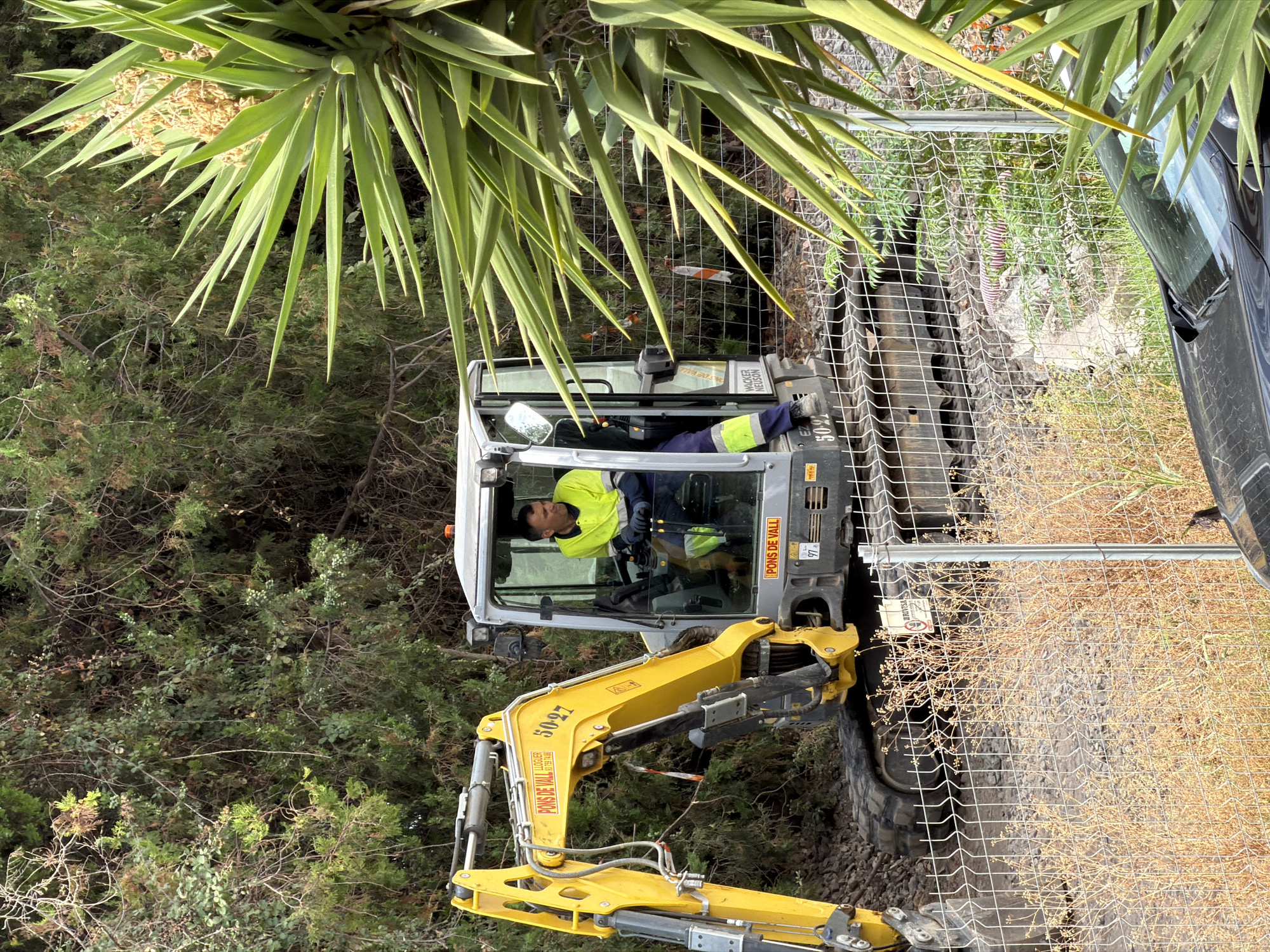 A male construction worker in a high-visibility yellow jacket and blue pants is operating a yellow and gray excavator. The excavator has "PONS DE VALL" and "LLOGUER 93 759 74 86" written on its arm, along with "50-27". The worker is seated inside the cab of the excavator, looking to the right. The scene appears to be in a construction area, with a wire fence in the foreground and dense foliage in the background. A portion of a black car is visible in the lower right corner of the image. The environment suggests an outdoor, possibly rural or suburban, location, likely in Salou, Spain, given the text on the excavator.