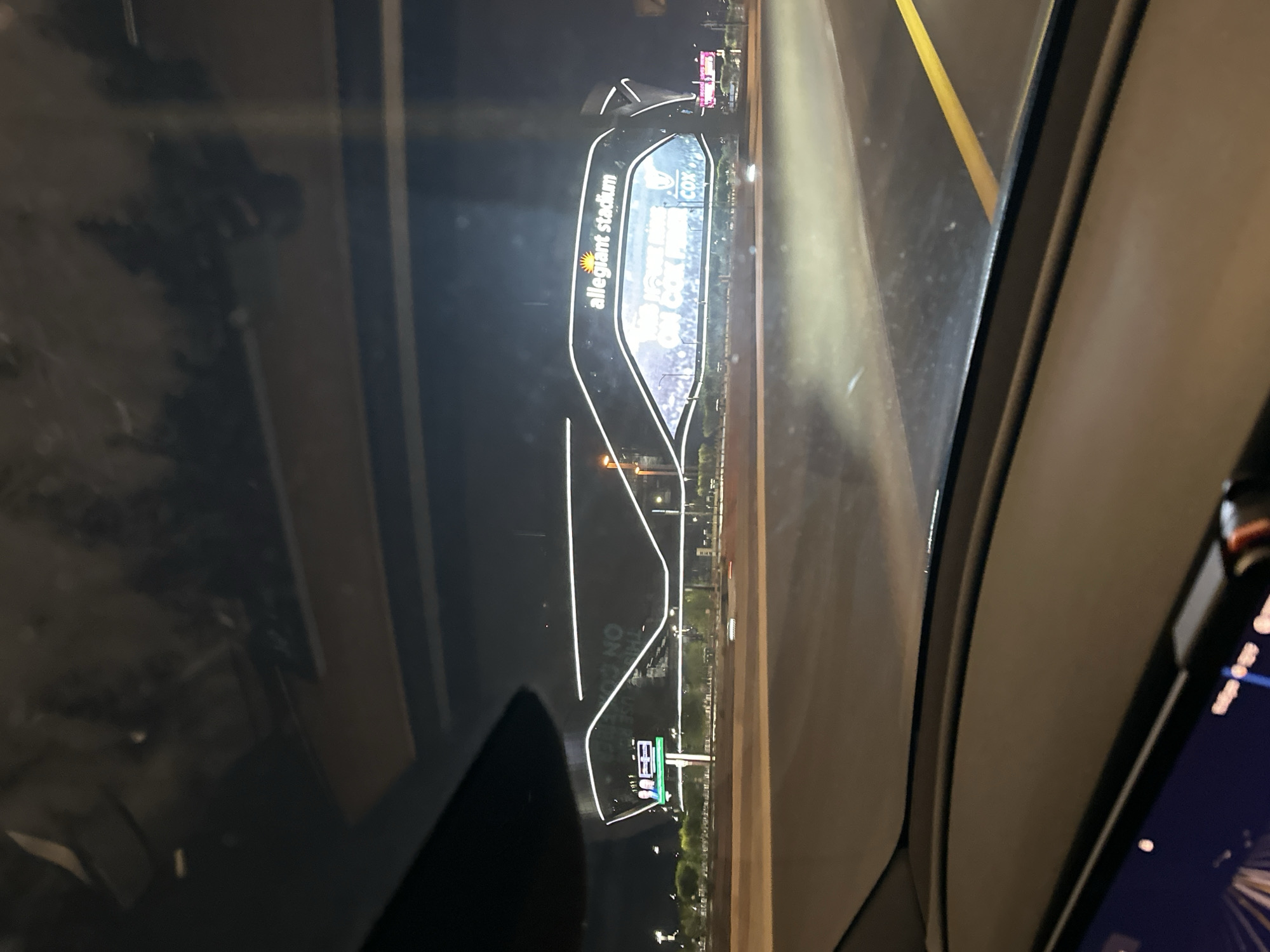 The image shows a nighttime view of Allegiant Stadium in Las Vegas, Nevada, as seen from a vehicle on a highway. The stadium is illuminated with white lights, and a large screen displays advertisements. The perspective is from the driver's seat, with the dashboard and windshield visible in the foreground. Trees and other lights are visible in the background, indicating an urban environment.