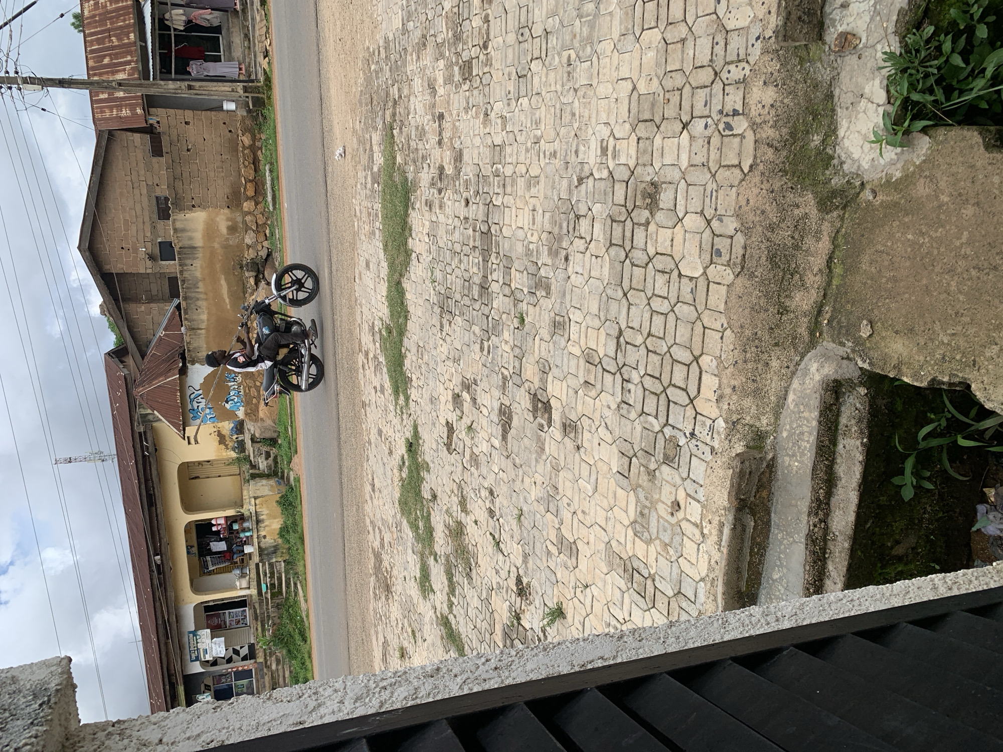 A man rides a motorcycle down a paved street in front of shops. The man is wearing a black cap, a white shirt with black sleeves, and dark pants. The motorcycle is black with silver accents. The shops are made of brick and stucco and have open fronts. There are clothes displayed in one of the shop windows. The street is lined with concrete pavers. The sky is cloudy.