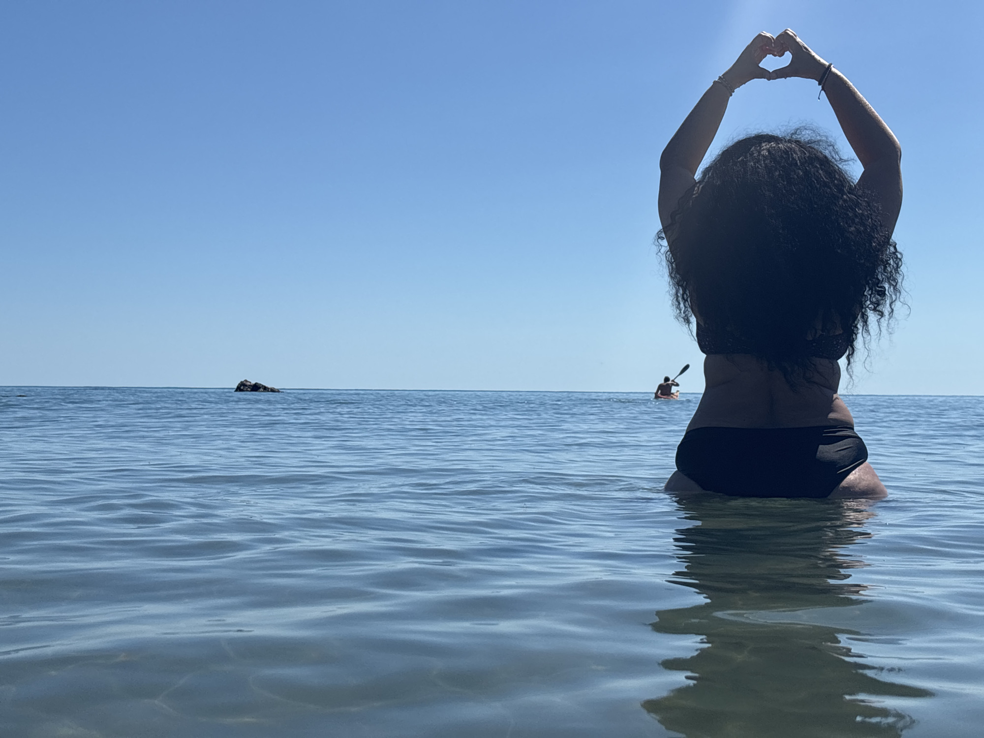 A person with long, dark, curly hair sits in the clear, blue water of Rezovo, Bulgaria. They are seen from behind, facing away from the camera, and are making a heart shape with their hands above their head. The person is wearing black bikini bottoms. In the distance, on the calm, rippling water, there are two kayaks with individuals paddling. A small rocky outcrop is visible on the horizon. The sky above is a clear, bright blue.