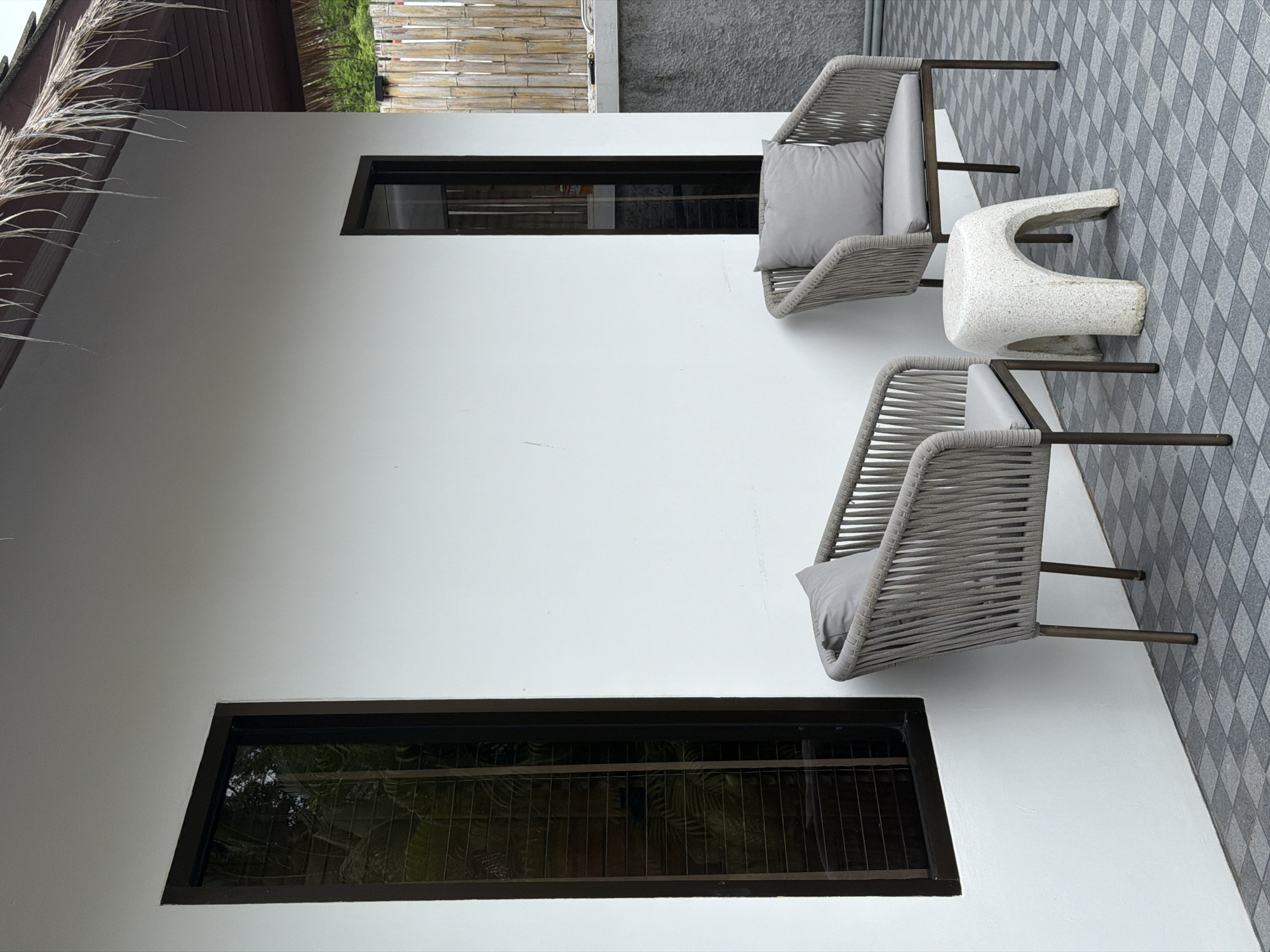 This is a full-shot, eye-level photograph of an outdoor seating area in front of a white stucco building in Baan Bo Phut, Thailand. The scene features two grey woven chairs with grey cushions and pillows, positioned on a grey checkered tiled patio. Between the chairs sits a small, white, textured stool with an arch-shaped base. To the left of the seating area, a tall, narrow window with a dark frame reflects some greenery. To the right, another window of the same size and frame is visible, and beyond that, a bamboo fence and a grey concrete wall are present. The building has a thatched roof overhang. No people are present in the image.