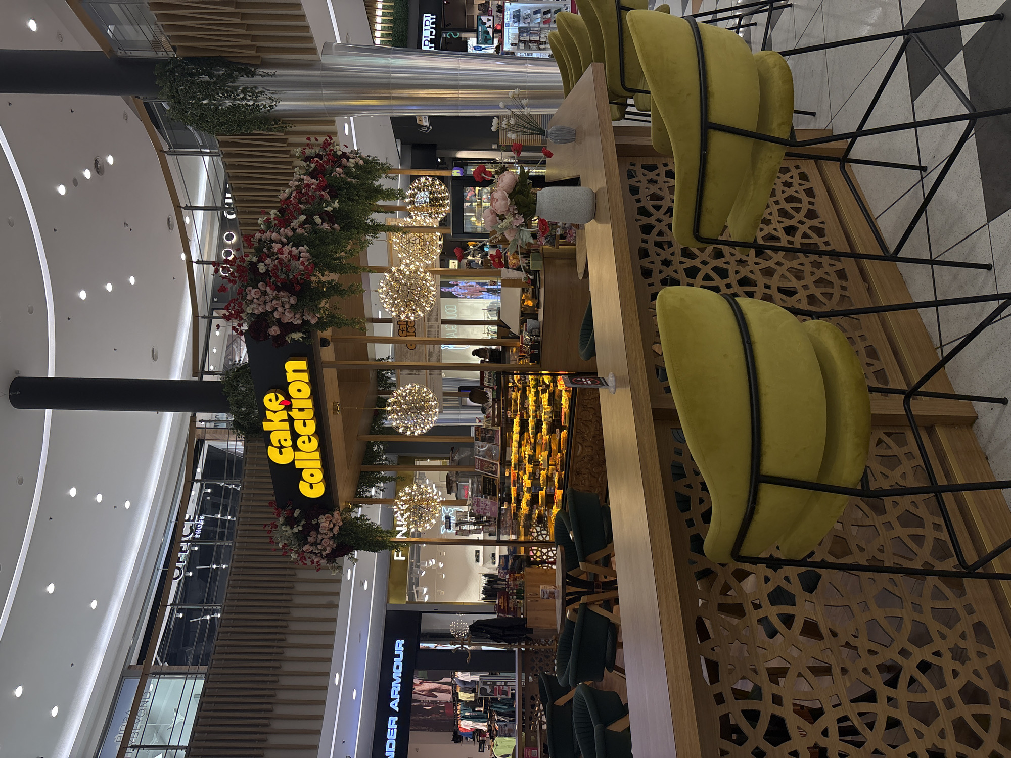 The image depicts the "Cake Collection" cafe in Plovdiv, Bulgaria, located inside a modern shopping mall. The cafe features a counter with yellow cushioned bar stools with black metal frames. Ornate wooden screens surround the cafe. The cafe is decorated with flower arrangements, spherical light fixtures, and greenery. The mall's interior includes clothing and shoe stores. The mall appears to have a multi-level structure. The scene is brightly lit, with a view extending across multiple shops and mall corridors. No people are immediately visible.