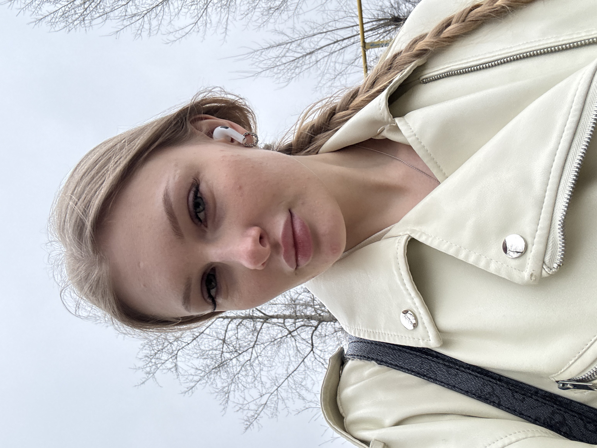 A young woman with fair skin and light blonde hair, partially braided, is wearing a white leather jacket and wireless earbuds in her left ear. She's looking down at the camera, likely taking a selfie. A black strap of a bag is visible across her body. The background shows bare tree branches against a cloudy sky, typical of late fall or winter. Given the location of Perm, Russia, it's plausible the photo was taken in a park or urban area where bare trees are common in the colder months.