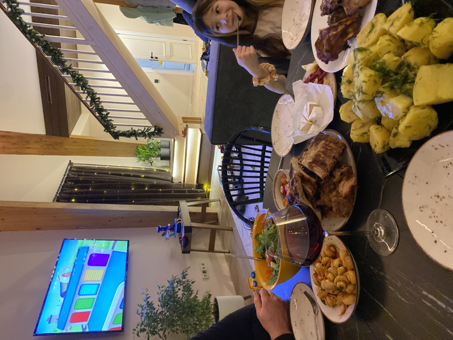 The image depicts an indoor dining scene occurring at a modern residential property in Burdakovka, Russia. The setting features light-toned wooden structural beams and a light-colored wooden staircase adorned with a green garland, indicating a possible holiday period. A large flat-screen television mounted on the left wall displays a colorful animated scene featuring a blue vehicle. Beneath the television, a small wooden console table holds a blue and white toy structure. Dark curtains with string lights are visible behind the console table, and potted plants are situated nearby and on a lighted step.
In the foreground, a dark-colored dining table is set with multiple dishes. Observable food items include a large plate of grilled ribs or meat, a plate of boiled potatoes with dill, a plate of white beans in a sauce, and a plate containing sliced cheese (likely Brie) and cured meat. A prominent orange bowl contains a green salad, and a wine glass filled with red wine stands on the table. Several empty or partially used white plates are also present.
Two individuals are partially visible. On the left, a person's arm and hand are near the salad bowl. On the right, a young girl with long, light brown hair, wearing a light-colored shirt, is shown from the chest up, holding a spoon to her mouth and appearing to be eating. She looks slightly off-camera with a subtle smile. A dark sofa is partially visible in the mid-ground on the right. The room is illuminated with warm artificial light.
