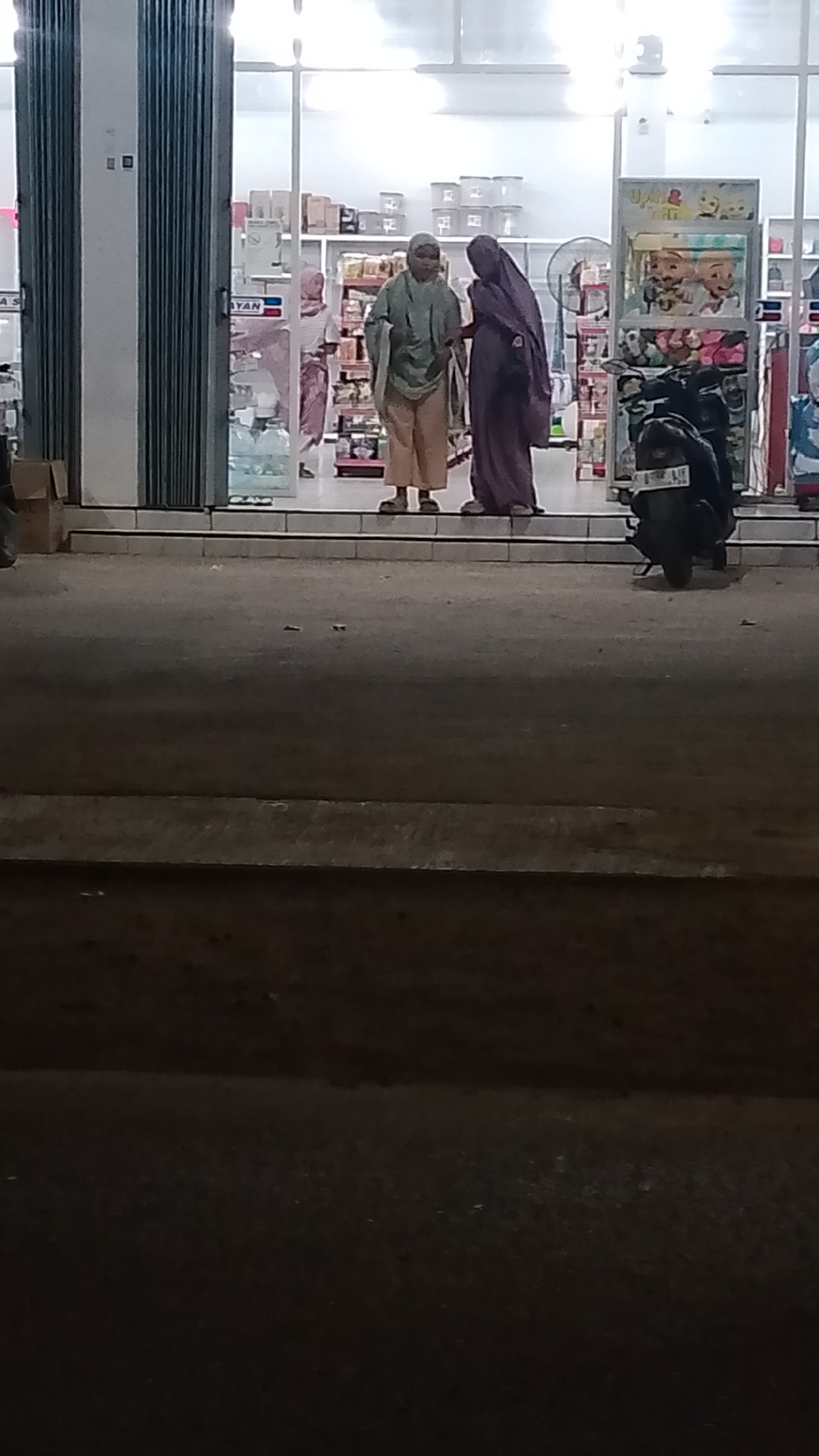 The image shows two women in traditional clothing standing on a sidewalk in front of a store. The store appears to be a convenience store, with shelves stocked with goods visible through the glass doors. A motorcycle is parked to the right of the store entrance. The scene is set at night, with the store lights illuminating the area. The women appear to be engaged in conversation. The overall impression is of a typical street scene in a residential or commercial area. Based on the context provided, the location is Karanglangu, Indonesia.