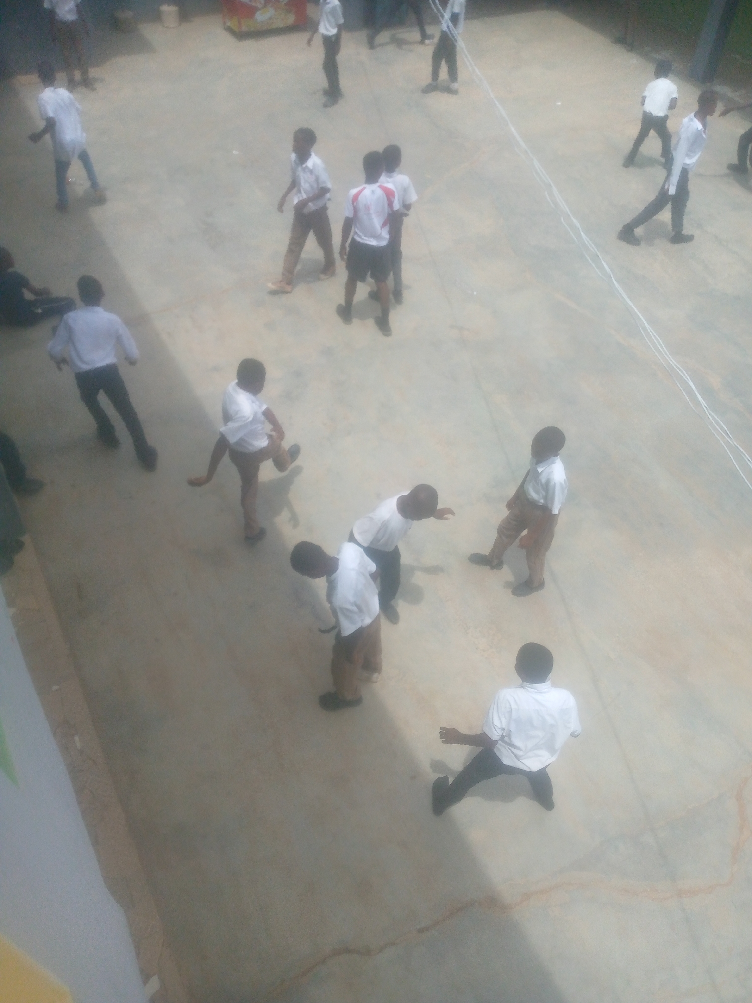 The image shows a group of students, primarily boys, gathered in an open, paved area, likely a schoolyard. They are dressed in school uniforms that consist of white shirts and varying colored trousers or shorts. Some students are standing in small groups, while others are moving around. The overall setting suggests a recess or break time at a school, somewhere in the city of Ikorodu, Nigeria.