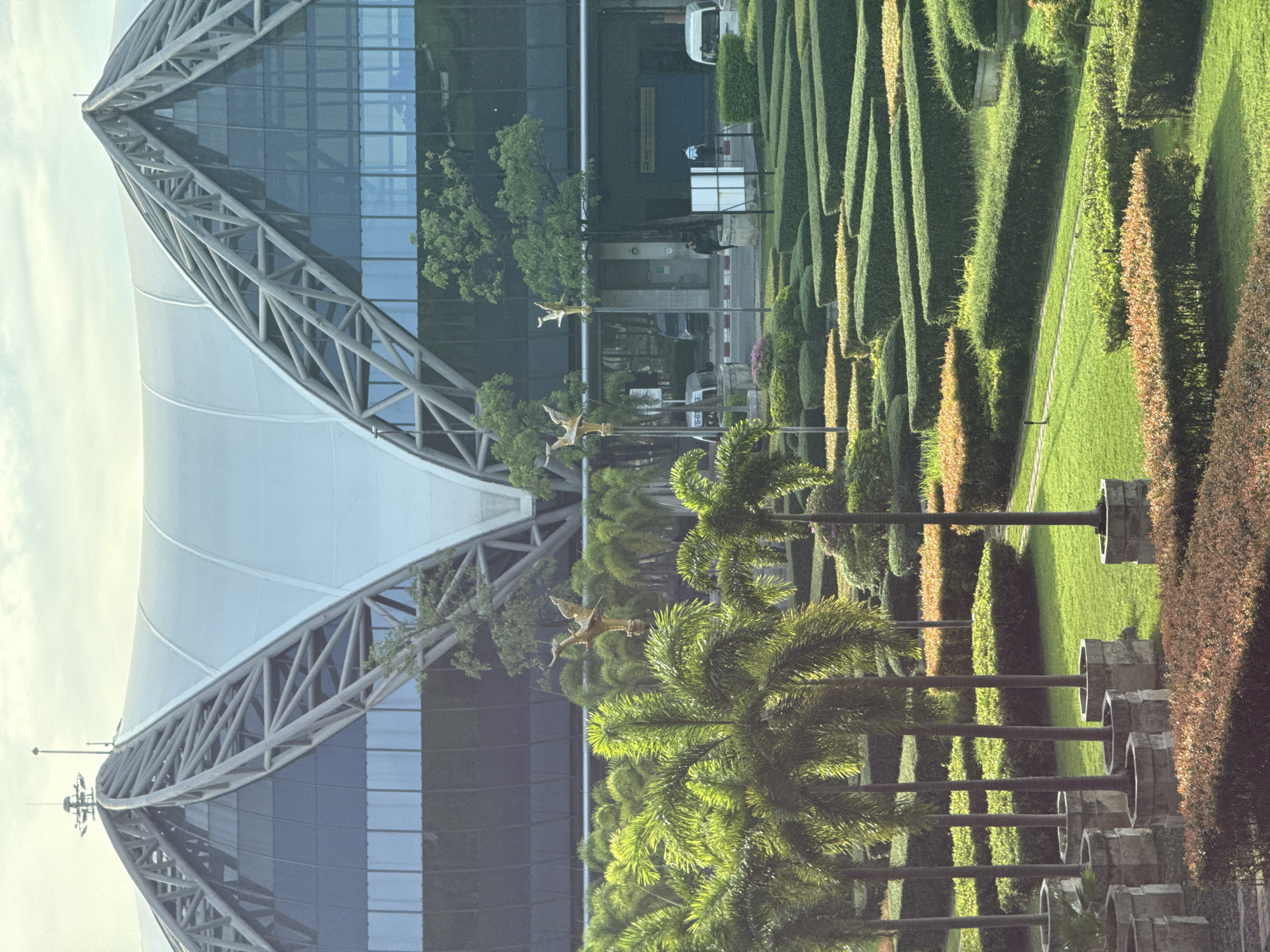 The image shows a view of Suvarnabhumi Airport in Nong Prue Subdistrict, Thailand. In the foreground, there is a meticulously landscaped garden with manicured hedges and palm trees in planters. The hedges are arranged in geometric patterns, creating a sense of order and depth. Golden bird statues are visible on poles amidst the foliage. In the background, the distinctive architectural structure of the airport terminal is prominent, characterized by its sweeping rooflines and glass facade. The sky is overcast, suggesting a diffused natural light. The scene depicts the airport's exterior, highlighting both its architectural grandeur and the surrounding, well-maintained grounds. There are a few vehicles and individuals visible near the terminal entrance.