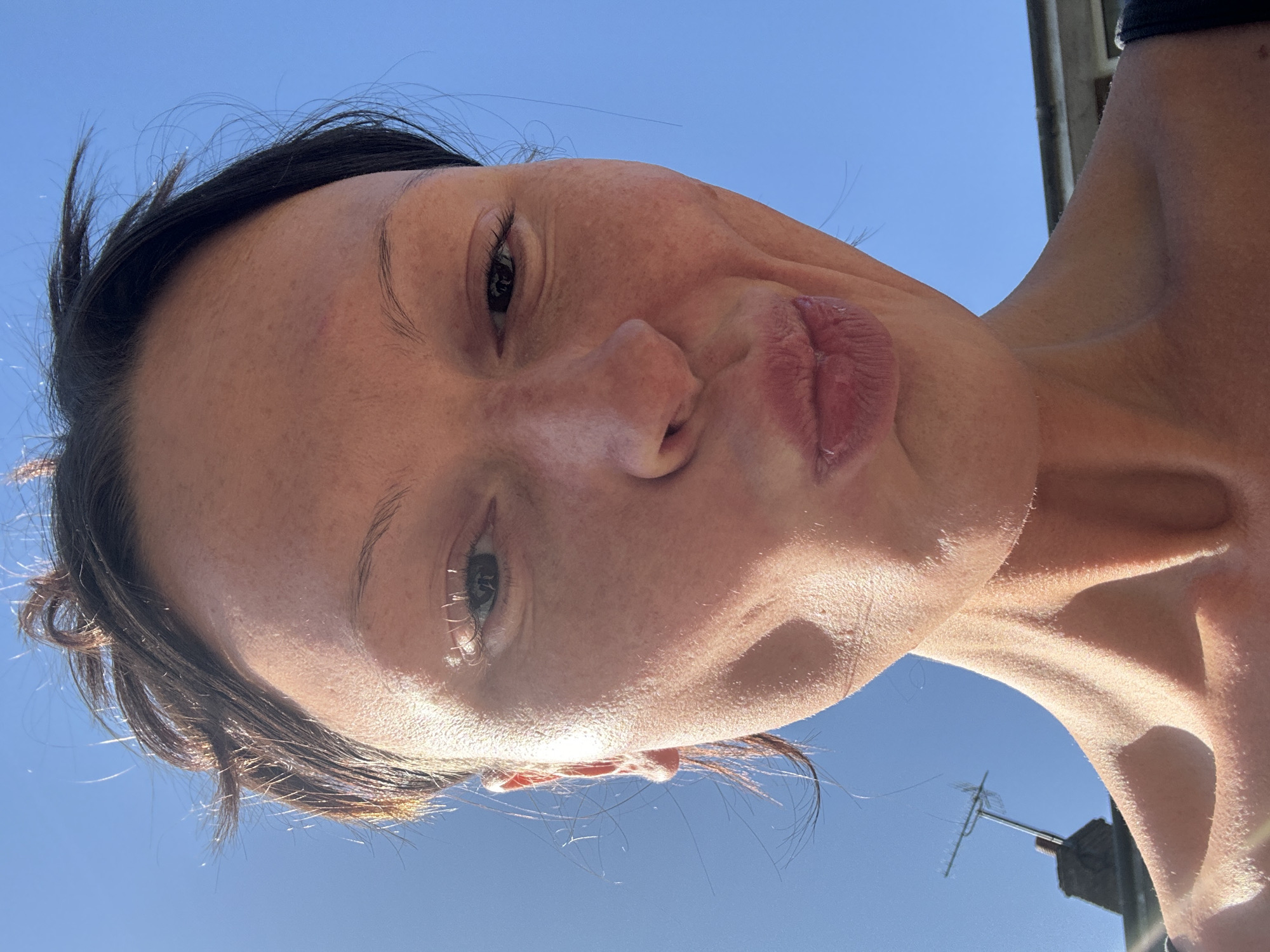 A close-up, low-angle portrait captures a person, likely female, looking towards the camera against a clear, vibrant blue sky. Her head and upper chest are visible, with dark hair framing her face. She has a playful expression, puckering her lips in a "kissing" pose, and her eyes show a slight squint, possibly from the bright sun. Strong, direct sunlight illuminates her features, creating prominent highlights on her skin and casting some shadows.
The setting is outdoors on a sunny day, indicated by the clear blue sky and the harsh, bright light. In the upper right background, a dark building or structure is partially visible. Towards the lower right, a television antenna or aerial rises against the sky, suggesting an urban or suburban environment within Tunbridge Wells, United Kingdom. The overall impression is a casual, lighthearted moment captured under a bright sky. There is no visible text in the image.