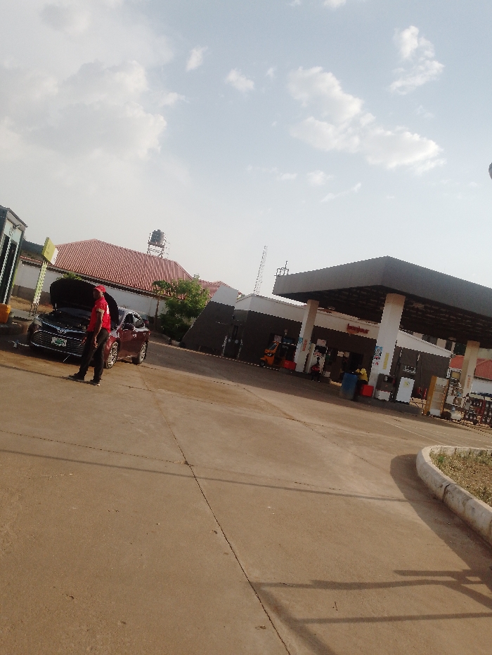 The scene depicts the forecourt of a Conoil gas station located in Sabon Tasha, Nigeria, under a bright, partly cloudy sky. The ground is a light-colored concrete surface. In the left midground, a dark sedan, likely maroon, is stationary with its hood raised. A male subject, wearing a red short-sleeved shirt, black trousers, and black shoes, stands adjacent to the driver's side of the sedan, facing the open engine compartment. To the far left, a fuel pump dispenser unit is visible. Behind the sedan and pump, a building with a terracotta-tiled roof features a black water tank on an elevated metal stand. To the right, the main Conoil station structure is present, characterized by a dark grey canopy supported by white columns. The Conoil logo is visible on the building facade. Underneath the canopy, multiple fuel pump dispensers are positioned. An orange three-wheeled vehicle is parked near these pumps. A second subject, wearing a yellow shirt, is discernible near a blue receptacle on the right side of the pumps. A concrete curb with sparse vegetation defines the right edge of the forecourt. A tall metal mast rises in the distant background.