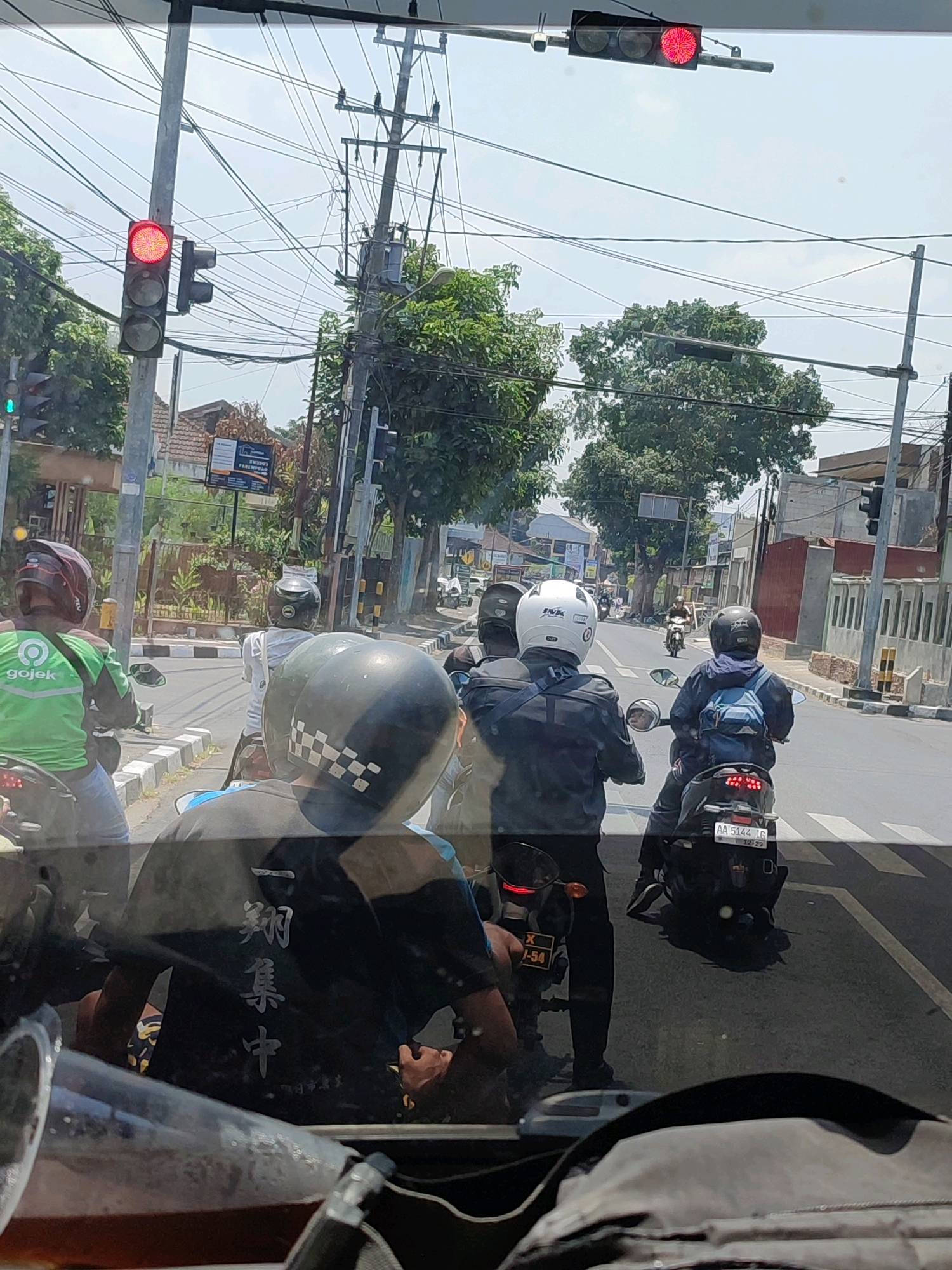 The image depicts a street scene in Magelang, Indonesia, during daylight hours. The perspective is from inside a vehicle, looking out at traffic. In the foreground, parts of the vehicle's interior, including a reflective surface and possibly a drink container, are visible.
The street is populated by several motorcyclists. One individual in the immediate foreground, in the lower center of the image, is wearing a black shirt with white Chinese characters printed vertically on the back and a checkered helmet. To their left and slightly behind is another motorcyclist wearing a green Gojek jacket. Several other motorcyclists in helmets and jackets are visible further down the road, some riding in a queue.
A traffic light is prominently displayed on a pole on the left side of the image, showing a red light. Another traffic signal is visible above and to the right, also illuminated red. The environment includes trees, buildings lining the street, utility poles with numerous electrical wires, and road markings including a pedestrian crossing. The sky is bright and overcast. The overall impression is of a busy urban intersection with traffic waiting at a red light.