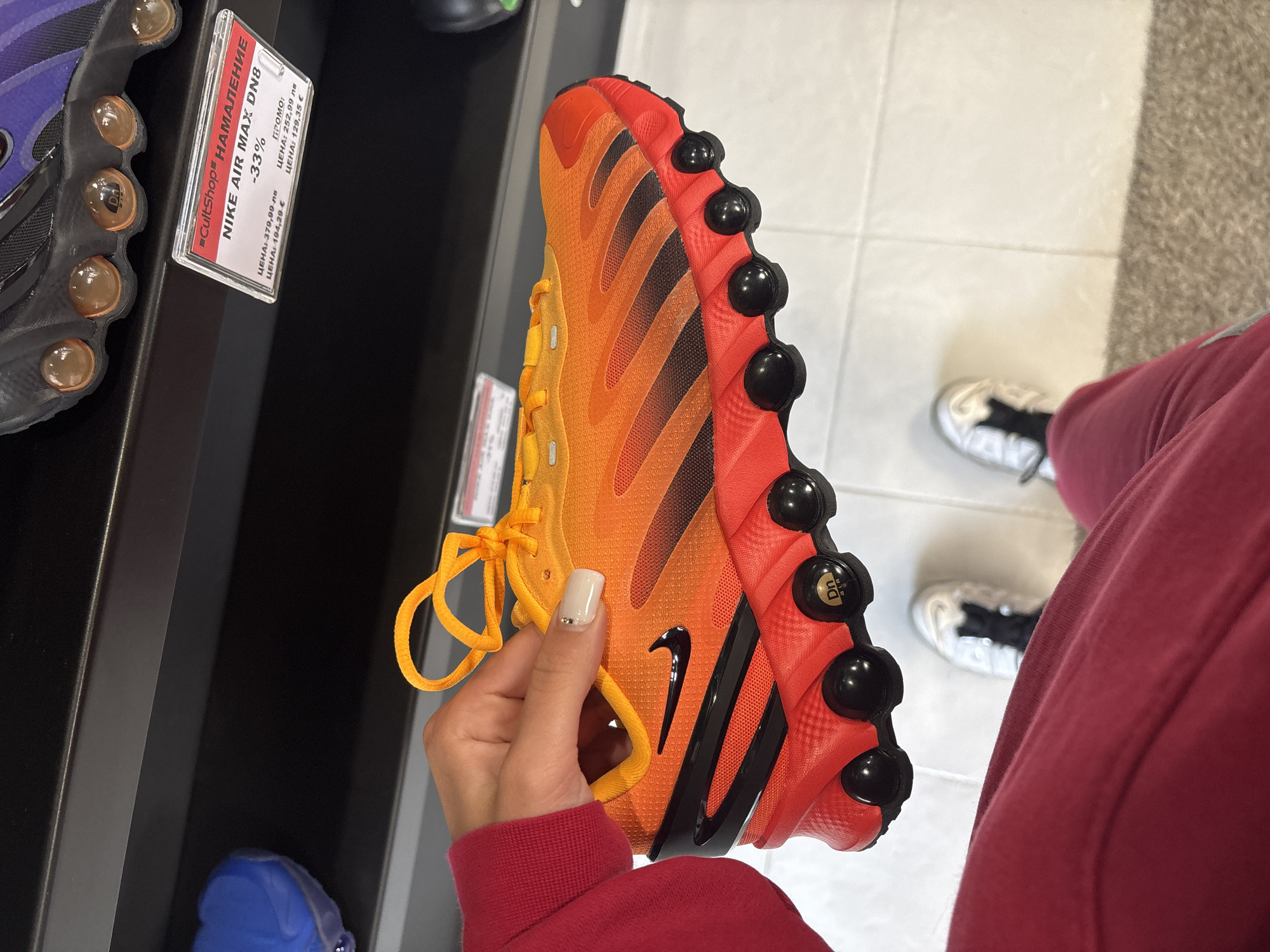 Here is a detailed description of the image:
**Subject:** The image focuses on a Nike Air Max Dn8 shoe, specifically an orange and red colorway, being held up by a person's hand.
**Environment:** The scene appears to be inside a retail store. A shoe display unit is visible behind the shoe, and tiled flooring is seen on the right side.
**People:** A person is holding the shoe. Only their hand and part of their red sweatshirt are visible. Their fingernail is polished with a light color. Another person is standing nearby, wearing shoes with black and white laces.
**Objects:**
* The Nike Air Max Dn8 shoe is the main object.
* A price tag is attached to the shoe display unit. The price tag mentions the name "CultShop" and the words "НАМАЛЕНИЕ" which is the Bulgarian word for sale.
* Another shoe in the background on display appears to be blue and purple.
**Actions:** The person is holding up the shoe, presumably to examine it or consider purchasing it.
**Location:** Given the text on the price tag, which is in Bulgarian, it is likely the store is in Bulgaria. Without additional information from the picture, it is hard to say where in the city of Gabrovo this picture was taken.