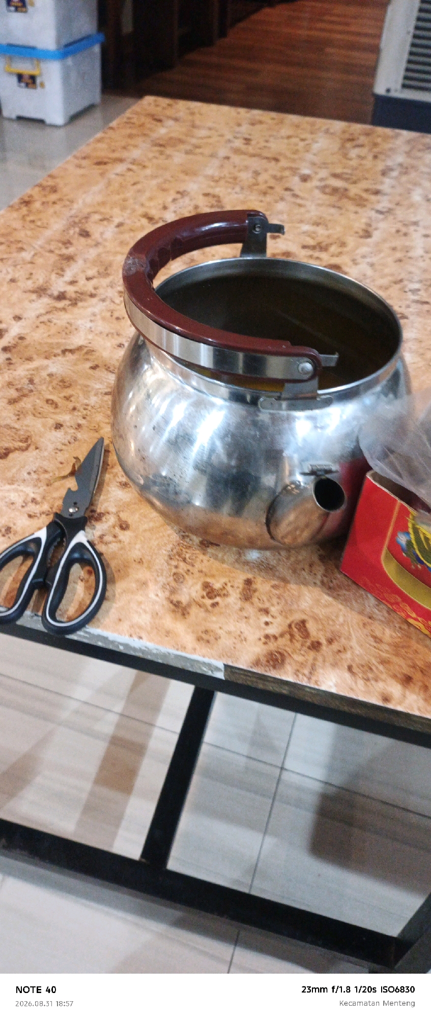 A close-up, eye-level shot shows a metallic kettle with a red handle on a table with a wood-grain patterned laminate surface. To the left of the kettle are black and white handled scissors. In the background, out of focus, are two white storage bins with blue lids, and a wooden floor. The lower right corner of the image shows the bottom edge of a red box with yellow accents. A black metal frame borders the bottom of the image, and text at the bottom reads "NOTE 40", "2026.08.31 18:57", "23mm f/1.8 1/20s ISO6830", and "Kecamatan Menteng". This last text indicates the location is Menteng District, Central Jakarta, Indonesia.