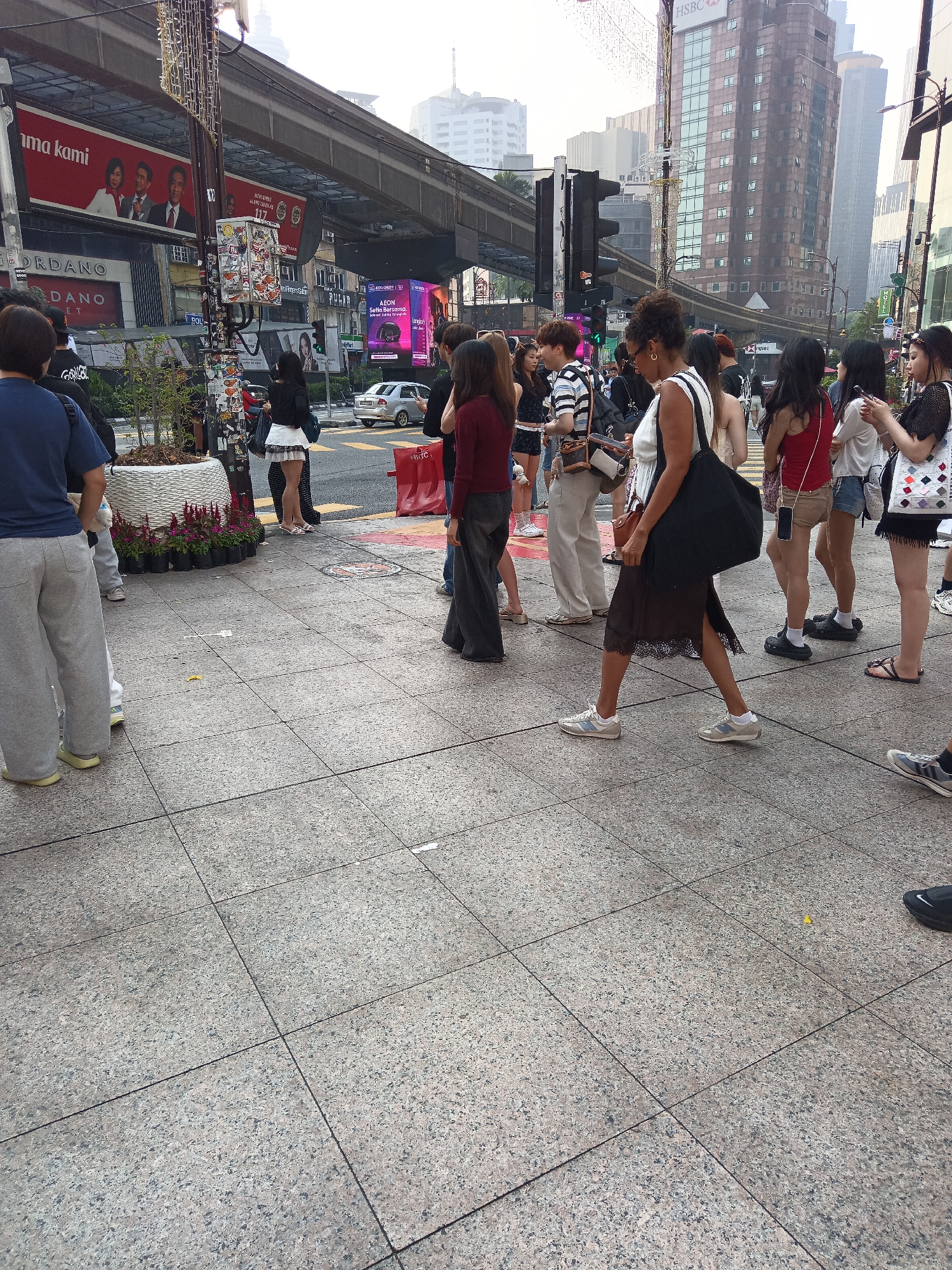 The image captures a bustling street scene in Kuala Lumpur, Malaysia, likely near the Bukit Bintang area, known for its commercial and entertainment hubs. In the foreground, a paved walkway with large, textured tiles is visible, with several people walking on it. A prominent feature is a large planter box filled with vibrant red flowers.
To the left, a person in a blue t-shirt and grey sweatpants is seen from behind. Further down the walkway, a group of individuals are gathered, some looking at their phones, while others stand or walk. The crowd includes several young women dressed in casual, fashionable attire, such as skirts and shorts, and some wearing sneakers.
The background reveals a typical urban landscape of Kuala Lumpur. Elevated train tracks, characteristic of the city's public transport system, run overhead. Several multi-story buildings are visible, including one with the HSBC logo. Advertisements are displayed on large billboards, with one featuring three people in formal wear. Traffic lights and street signs indicate an active intersection. The overall atmosphere suggests a busy commercial district during daylight hours.