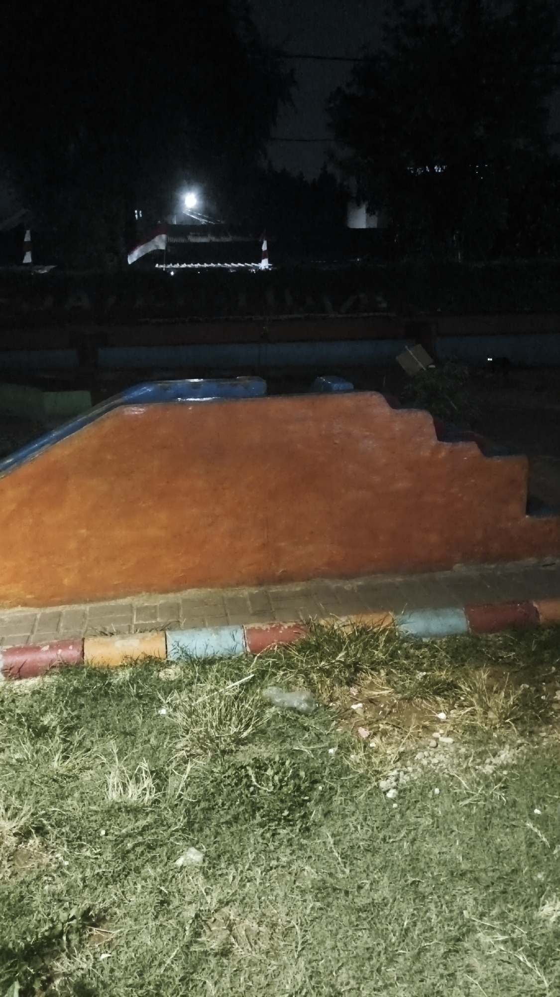 This image depicts a brightly colored playground structure at night, located in a grassy area. The structure itself is predominantly orange with blue accents along the top edge, suggesting it may be a slide or a climbing apparatus with steps. The base of the structure is bordered by a segmented curb painted in alternating colors of red, yellow, and blue. Beyond the playground equipment, a dark background suggests trees and some distant structures, possibly buildings or rooftops, illuminated by a bright light source. The overall scene is dimly lit, consistent with nighttime photography. The location is identified as South Jakarta, Indonesia. There are no people or animals visible in the frame, and the environment appears to be a public outdoor space, likely a park or playground.
