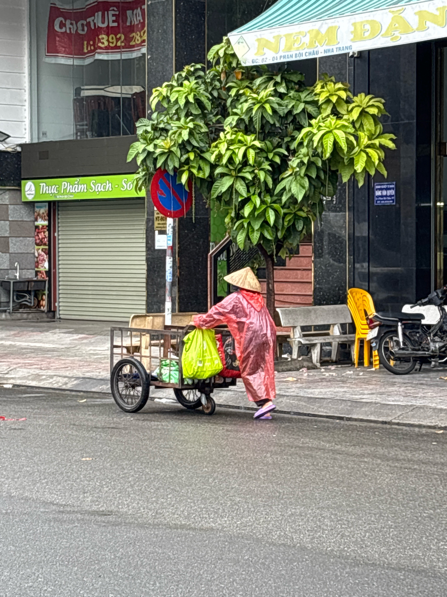 The image depicts a street scene in Nha Trang, Vietnam, on a wet day. A person wearing a red raincoat and a conical hat is pushing a metal cart laden with green bags and some boxed items. The cart has two large wheels and a smaller swivel wheel at the front. In the background, there is a building with a green awning that reads "Thực Phẩm Sạch - Sổ" and another sign above it with Vietnamese text. Adjacent to this is a rolled-down metal garage door. A "no parking" sign is visible on a pole. Further to the right, a yellow plastic chair and a motorcycle are parked near a concrete bench. The ground is wet, indicating recent rain.