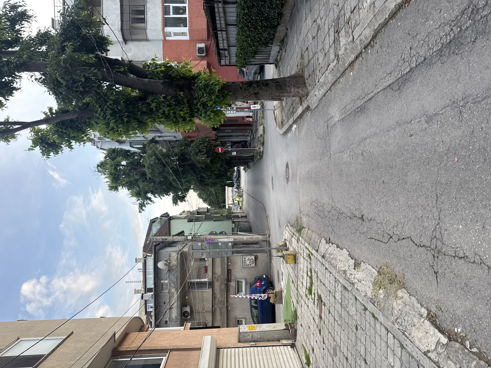 The image captures a static, sunlit view down a narrow, sloping urban street in Varna, Bulgaria, lined with multi-story residential buildings. The asphalt road shows signs of wear and cracking, and a cobblestone-patterned sidewalk with visible weeds runs along the right side.
On the left, several mature trees with full green foliage provide shade, obscuring parts of the buildings. Behind them, various buildings are visible: a light brown/tan structure in the foreground, followed by a gray/light green building featuring a satellite dish, multiple air conditioning units, and crisscrossing power lines. Further down, buildings with red/orange and white facades can be seen. On the right, a red/orange building with white window frames and another air conditioner is present, with a dense green hedge covering a portion of its lower wall.
A dark blue car, possibly a hatchback, is parked on the left side of the street, partially behind a temporary red and white striped barrier. This barrier has faint, blurry text, which appears to include "BEPEH" and "ЦЕНТЪР" (suggesting "Varna Center" in Cyrillic). A red circular "No Entry" traffic sign (a red circle with a white horizontal bar) is mounted on a pole near the blue car, amidst other less discernible signs. A yellow electrical box stands on the right sidewalk.
The sky is partly cloudy with patches of clear blue. The overall impression is one of a typical, lived-in Eastern European residential street, with no people or specific activities depicted. The time of day appears to be daytime, likely morning or afternoon, given the clear lighting.