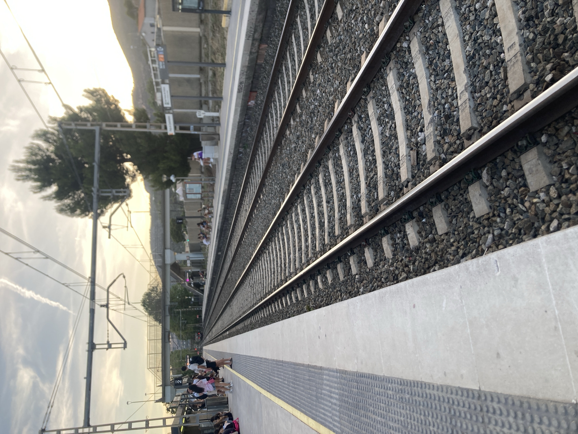 This is an eye-level shot of train tracks at the Castelldefels train station in Castelldefels, Spain. The tracks are on the right side of the image, with a platform on the left. The platform has a yellow line along the edge, followed by a grey, textured surface, and then a concrete platform. There are several people standing and sitting on the platform, some of whom are dressed casually. Overhead, there are power lines and structures for the train. In the background, there is a bridge or overpass, trees, and a hilly landscape under a partly cloudy sky with the sun setting.