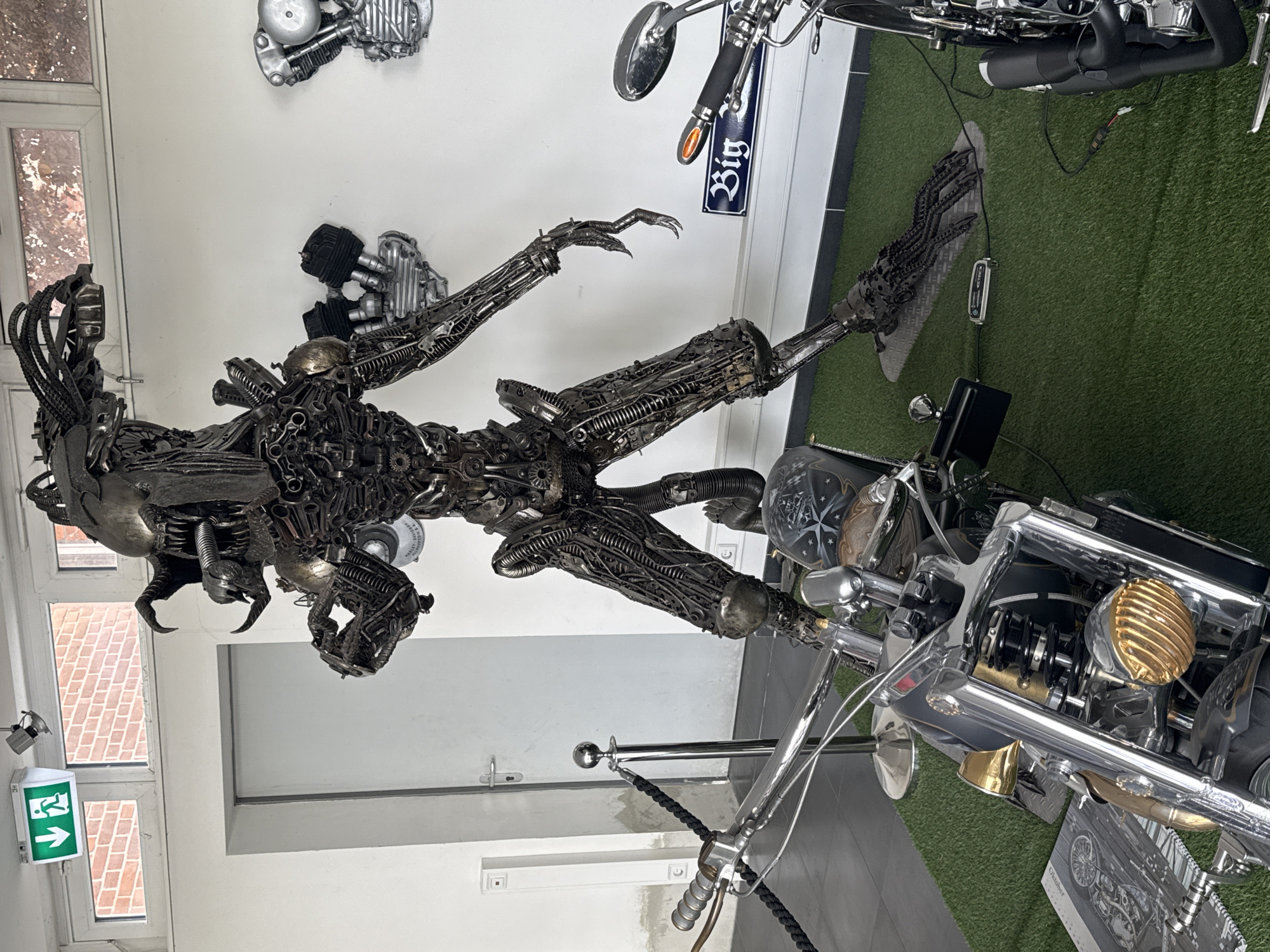 The scene depicts an indoor space with an eclectic collection of motorcycle-related items. Dominating the foreground is a large, metallic sculpture of an alien-like figure, constructed from various motorcycle parts and metal scraps. The sculpture is placed between two motorcycles, one positioned to the right and another partially visible above the sculpture. A "Big" sign is visible on one of the motorcycles. The floor is partly covered with green artificial turf. In the background, mounted on the white walls, are motorcycle engine components. Windows in the background show a brick building exterior and an exit sign. The scene appears to be in a showroom or a private collection. While Bietigheim-Bissingen, Germany, is specified, this information is not clearly visible in the image.