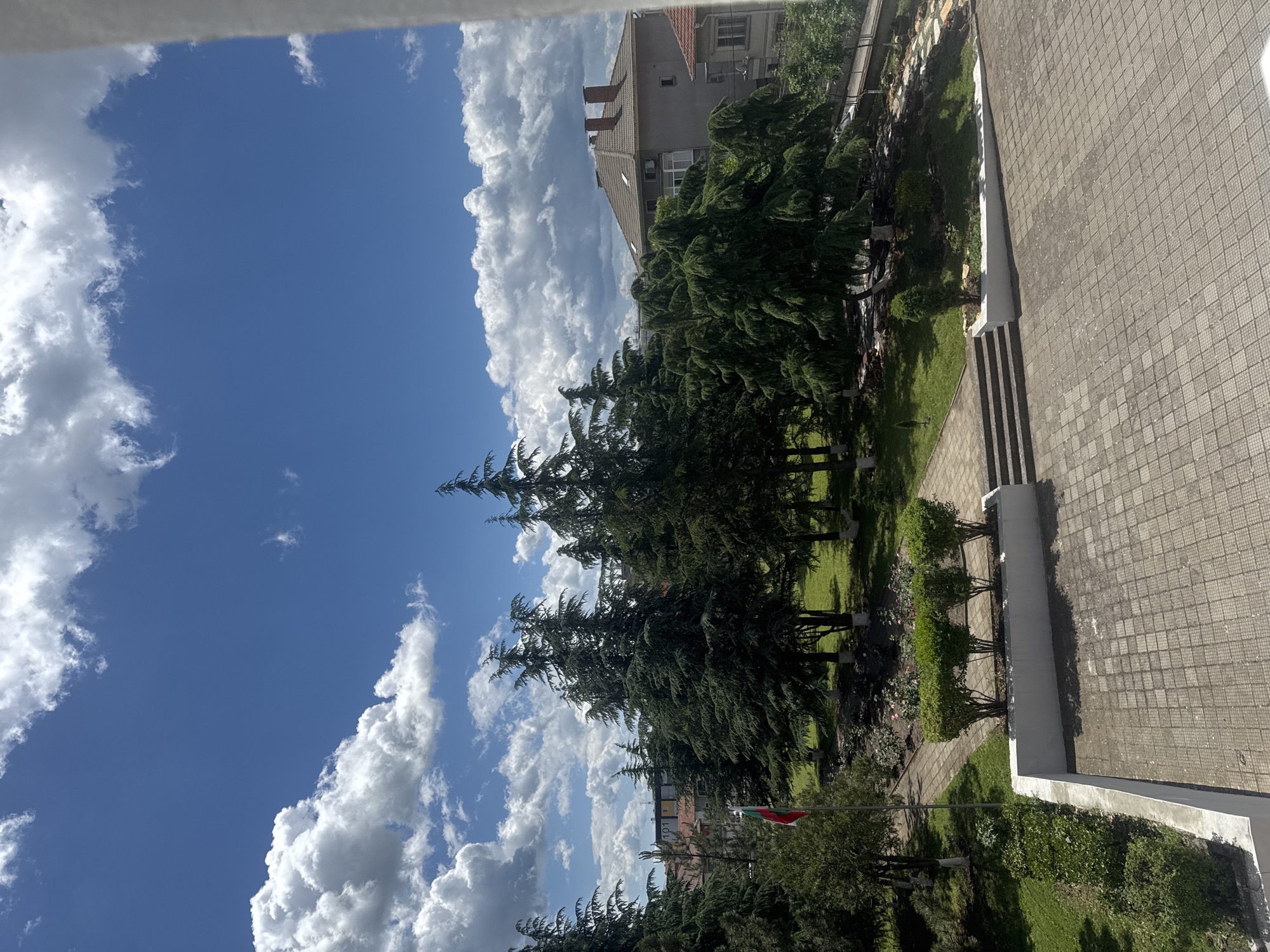 The image shows a view from an elevated position, likely a window, in Elhovo, Bulgaria. A blue sky with scattered white clouds dominates the upper left portion of the frame. A line of tall evergreen trees is centered, creating a visual division. To the right, there is a paved area with a set of steps leading to a building. A well-maintained garden area, with shrubs and a Bulgarian flag, is situated between the trees and the paved section. The background reveals buildings typical of a residential area. The composition suggests a peaceful urban landscape. There are no people in the visible area.