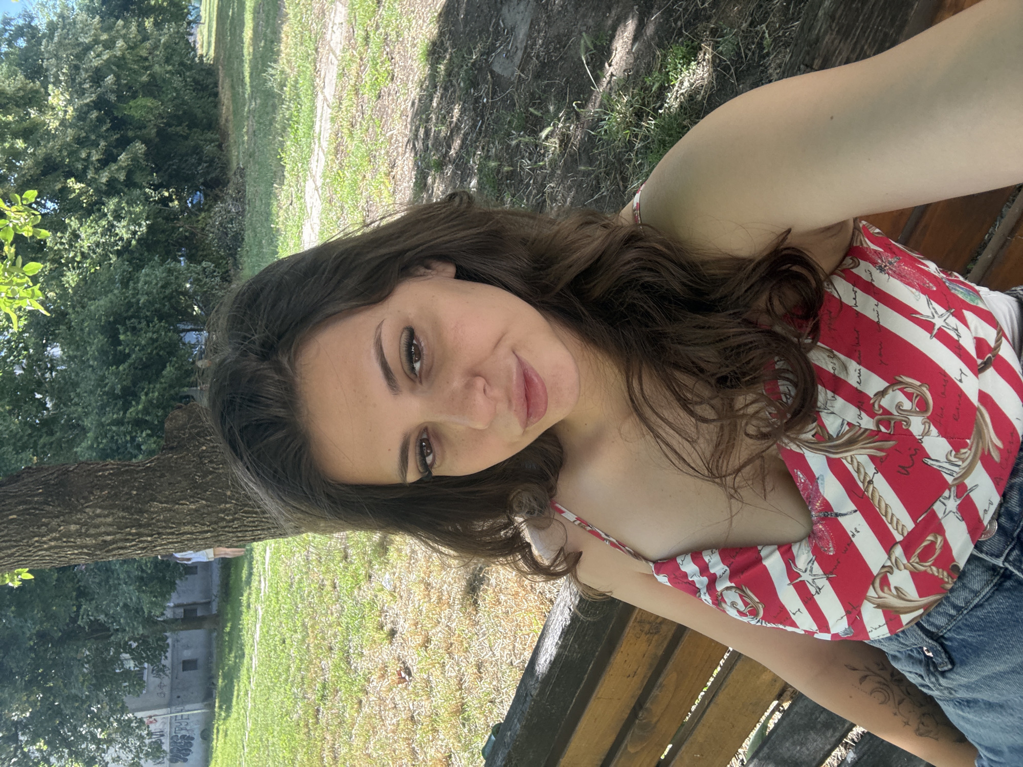 A young woman with long, dark, wavy hair is sitting on a wooden park bench, taking a selfie. She is wearing a red and white striped tank top with a nautical pattern, and blue denim shorts. Her left arm, which is visible, has a tattoo. She is looking directly at the camera with a slight smile. The background shows a park with green grass, trees, and a building in the distance. The location is Tsentralen Park in Plovdiv, Bulgaria, on a sunny day.