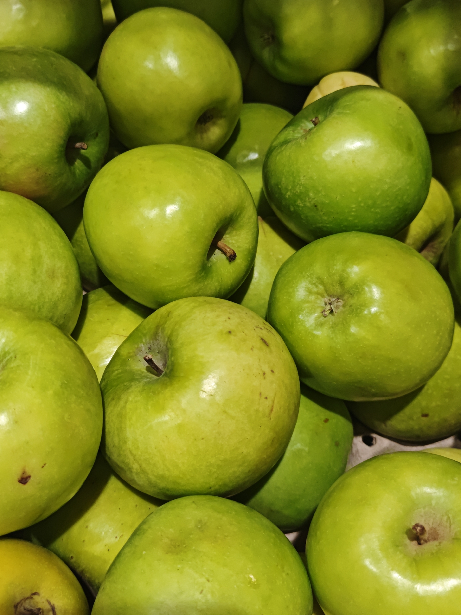 This media file is a close-up, top-down shot of a pile of bright green apples. The apples are round with some slight variations in shape and color, ranging from a vibrant lime green to a slightly yellower hue. Some apples display small brown blemishes or specks. The stems are visible on a few of the apples. The lighting is bright, highlighting the smooth, shiny skin of the fruit, and casting subtle shadows between them, suggesting an indoor setting like a market stall or grocery store display. There are no people, text, or specific location cues other than the provided context. The image focuses solely on the texture and color of the apples, creating a visually appealing arrangement of the fruit.