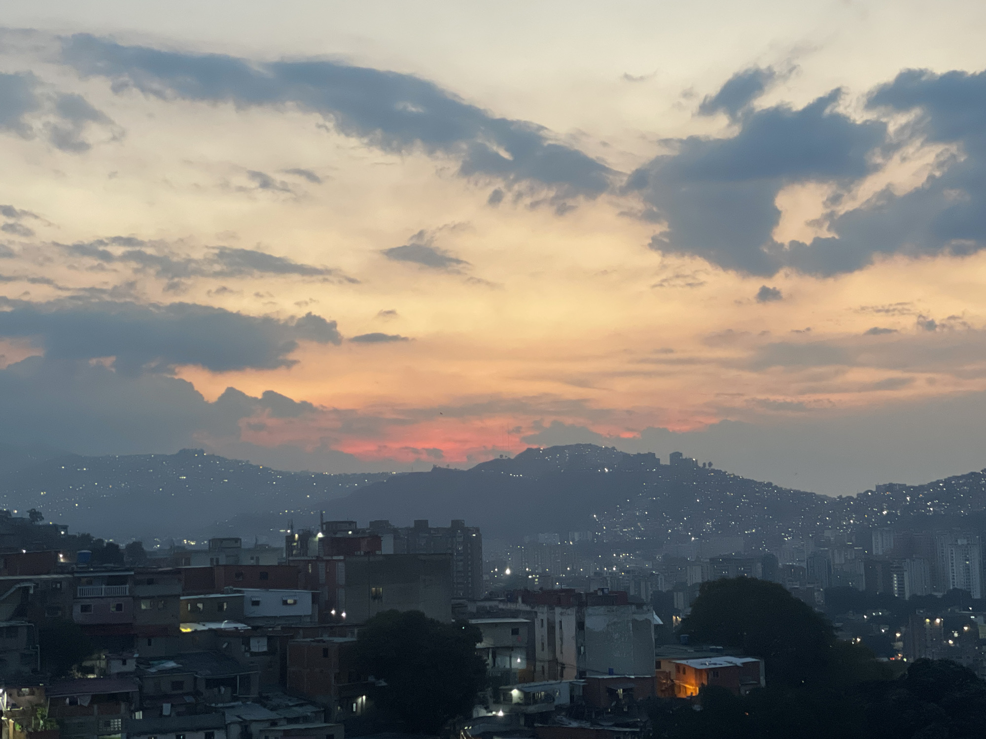 The image depicts a cityscape at dusk, likely Caracas, Venezuela, based on the mountainous terrain and urban density. The sky is a mix of orange, yellow, and grey hues, with clouds scattered across. Below the skyline, mountains are visible, covered with small, scattered lights indicating residential areas. The foreground shows densely packed buildings of varying heights and construction materials, with some streetlights visible. The overall scene is one of urban life against a backdrop of dramatic natural beauty.
