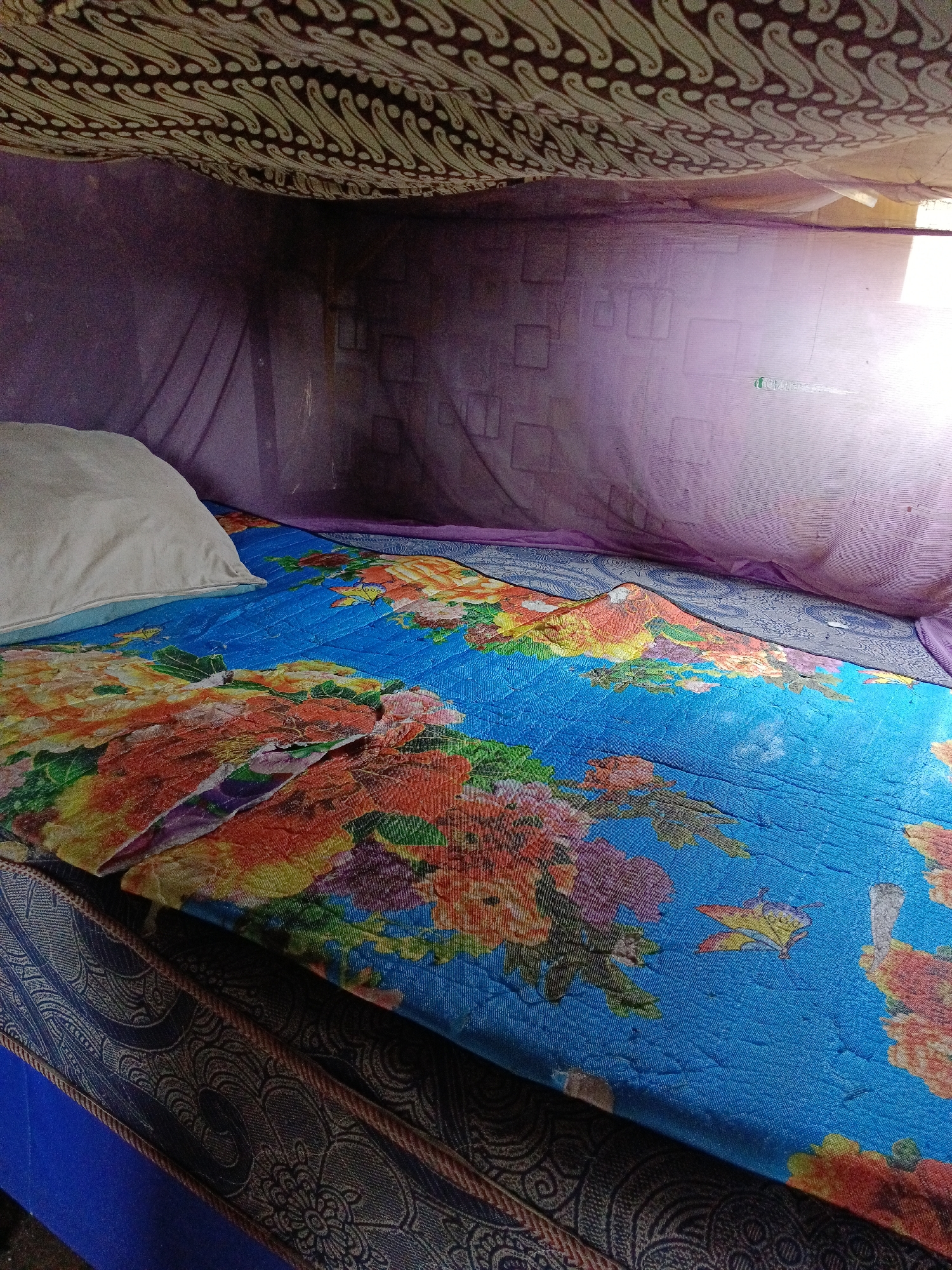 The image shows a close-up view of a bed, likely in a bedroom, with a floral patterned bedsheet. The bed is covered by a mosquito net, indicated by the sheer purple fabric draped over it. Above the bed, a patterned textile, possibly a batik fabric, is visible, suggesting it's part of a canopy or decorative element. The background shows a wall with a faint geometric pattern, and a window with natural light streaming in, creating shadows and highlights. The scene is set indoors, and based on the specific mention of the city of Aek Kanopan, Indonesia, this is a bedroom located in that region. There are no people or specific actions depicted in the image.