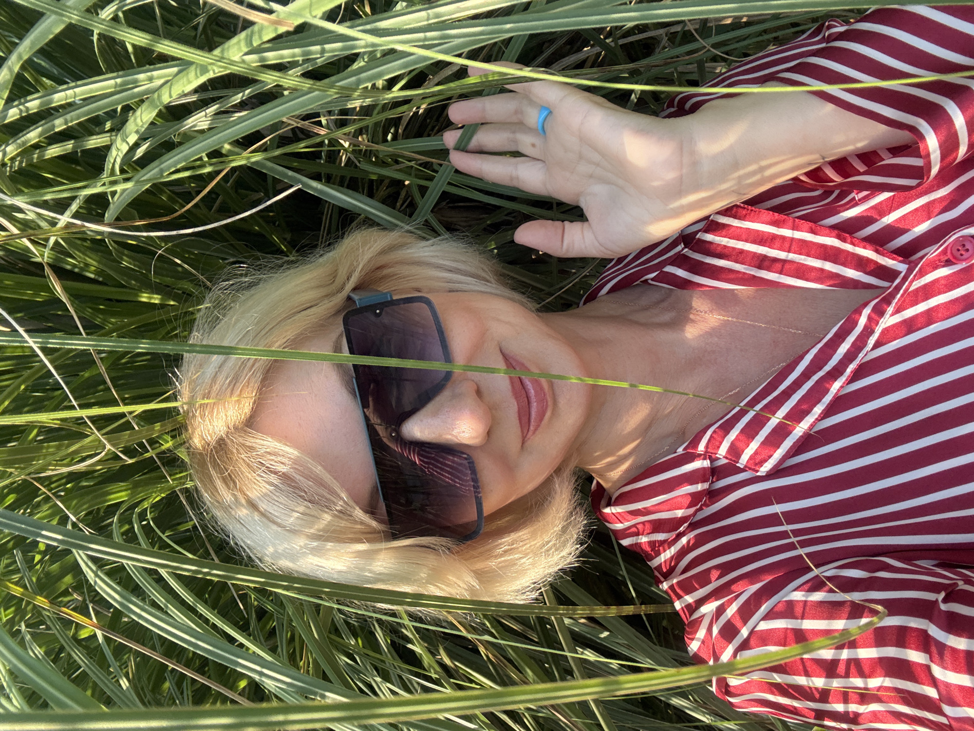 A medium shot captures a Caucasian woman with blonde hair and fair skin posing in tall green grass under sunlight. She is wearing large, dark sunglasses, a red and white striped shirt, and a blue ring on her right hand. Her right hand is raised, fingers spread, as if waving. The background is filled with lush, green foliage, suggesting an outdoor setting, possibly in a park or garden. The lighting indicates it is daytime, with shadows cast by the grass on the woman's face and hand. The overall scene is bright and natural.