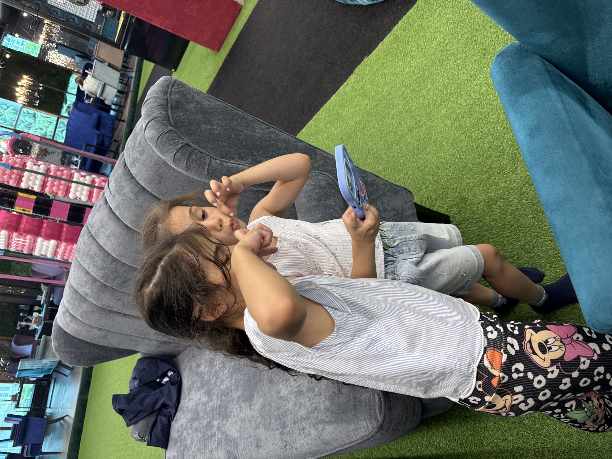 The image depicts two young girls interacting with a tablet computer, positioned in a brightly lit indoor space with a faux-grass floor. One girl, on the left, wears a striped blue and white top and patterned pants featuring Mickey Mouse. She has her arm extended towards her face, and her eyes are closed. The other girl, on the right, wears a white sleeveless top and denim shorts. She holds the tablet in her right hand, her fingers are visible on the screen. She appears to be looking at the tablet.
The girls are seated next to a large, plush gray sofa with distinctive segmented backrest. In the background, other seating areas are visible, including blue upholstered chairs and booths. The overall ambiance suggests a commercial or entertainment venue, possibly a cafe or an indoor play area. The flooring transitions from green turf to black carpet in sections. Further in the background, decorative elements like strings of lights and what appear to be illuminated panels are present, adding to the artificial environment. The area is well-lit, with artificial lighting contributing to the bright atmosphere. The presence of tables and chairs in the background indicates a public space designed for patrons. Given the context of the prompt, this location is identified as being in the city of Sochi, Russia.