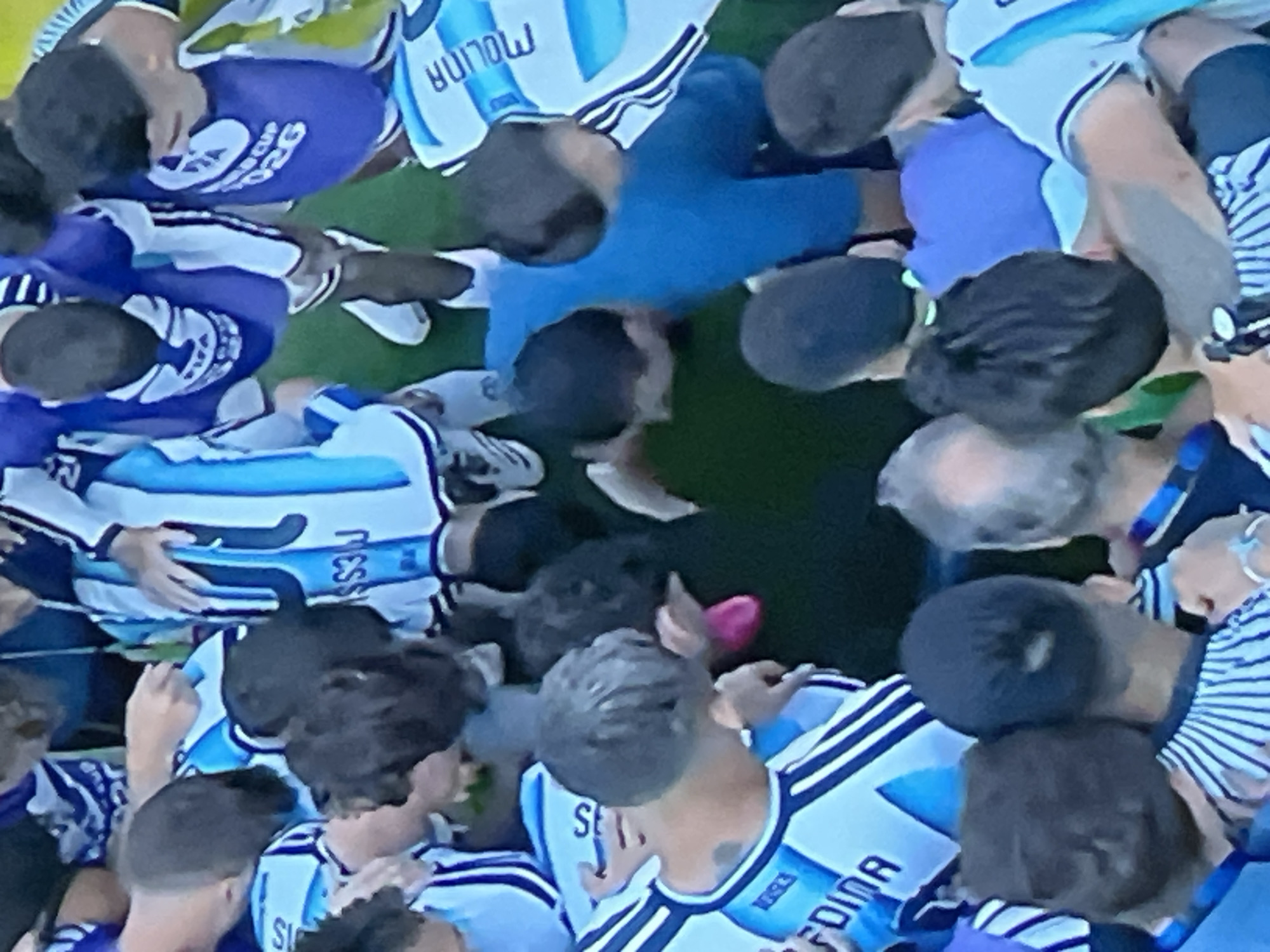 The image shows a group of men, likely soccer players, huddled together in a circle. They are wearing blue and white jerseys with stripes and some have the FIFA World Cup logo visible. One man in a blue shirt is bent over, possibly in a huddle or receiving attention. The ground appears to be a green surface, possibly grass or turf, suggesting an outdoor sports field. The lighting indicates it is daytime. The scene captures a moment of intense interaction or celebration among the individuals. The specific location in Jos, Nigeria is not discernible from the provided image.