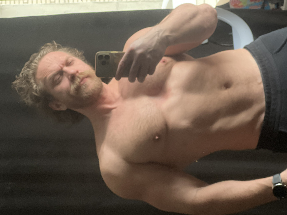 An adult male with fair skin, light brown curly hair, and a prominent mustache and beard is depicted bare-chested, taking a mirror selfie indoors in the city of Saint Petersburg, Russia. He wears black sweatpants and a black smartwatch on his left wrist. The man holds a smartphone, likely an iPhone with multiple rear cameras, in his right hand to capture the reflection. His facial expression is a grimace, with squinted eyes and furrowed brows. The background primarily features a dark curtain or wall, with a window frame and a partial circular light source, possibly a ring light, visible on the right.