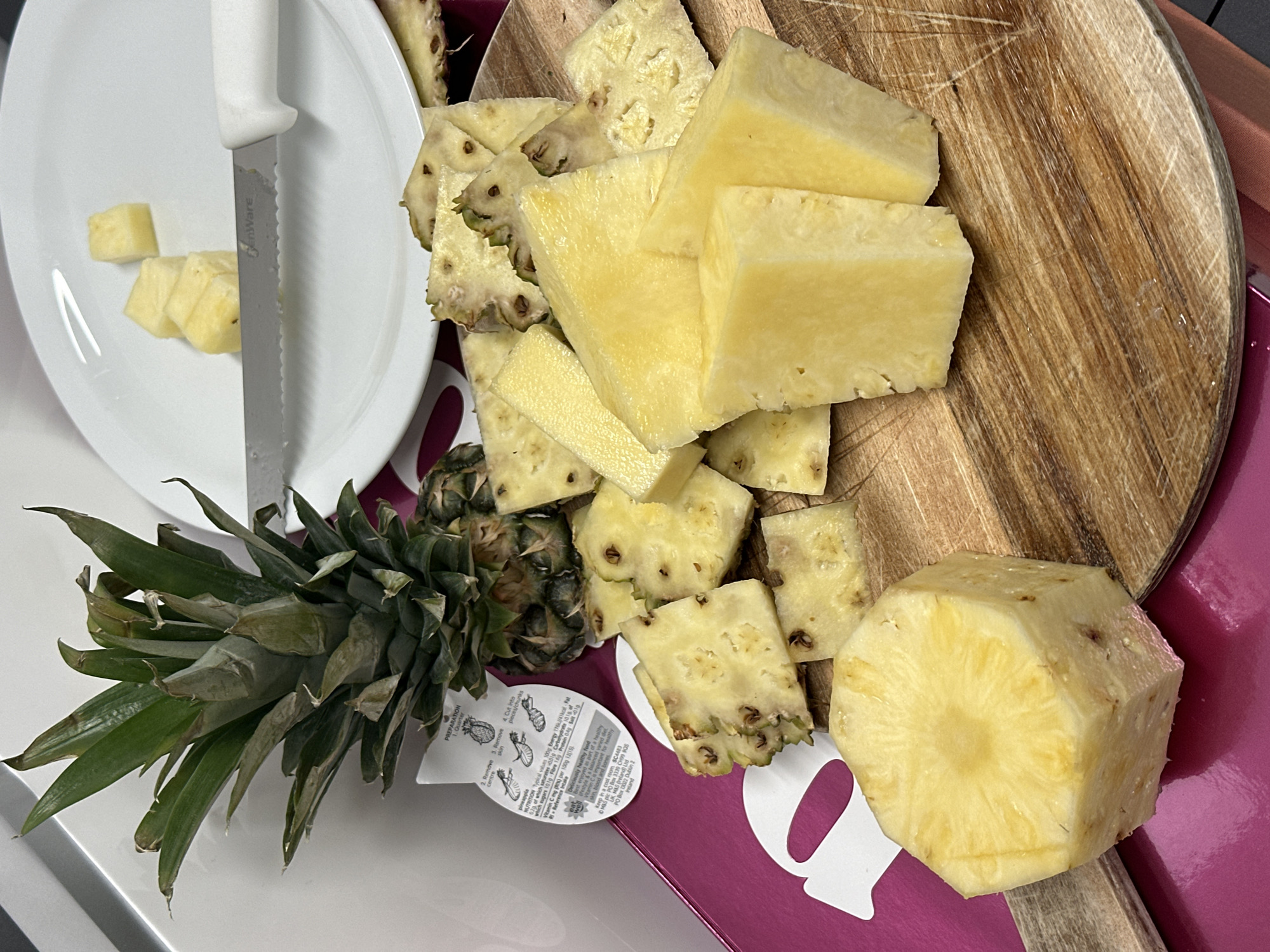 A partially-prepared pineapple sits on a white surface and a wooden cutting board with a white plate. The pineapple is in various states of preparation: a whole, peeled portion; chunks with the skin partially removed; and smaller pieces cut into cubes on the plate. A white knife rests on the plate, partially obscured by the pineapple cubes. A label with instructions for preparing the pineapple is visible near the uncut portion of the fruit. The background includes a pink-colored surface.
