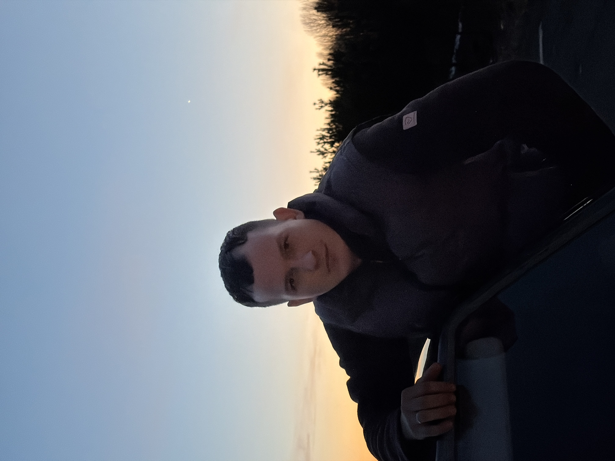 The image features a young man leaning out of what appears to be a car window, looking directly at the camera. The setting is outdoors during either sunset or sunrise, evidenced by the sky displaying a gentle gradient from pale blue at the top to warm orange and yellow near the horizon. A faint crescent moon is visible in the upper blue sky.
Silhouetted bare trees are discernable along the distant horizon, suggesting a rural or natural landscape. The man has short, dark hair and a neutral expression. He is wearing a dark-colored, possibly grey or black, padded jacket. A small, light-colored square logo or patch is visible on his left shoulder area. His right hand rests on the dark frame of the car window, while his left hand can be seen in a cropped view gripping an interior car handle, confirming the vehicle context. The lighting is subdued, emphasizing the twilight atmosphere. This scene is set in Kharnavy, Russia.
