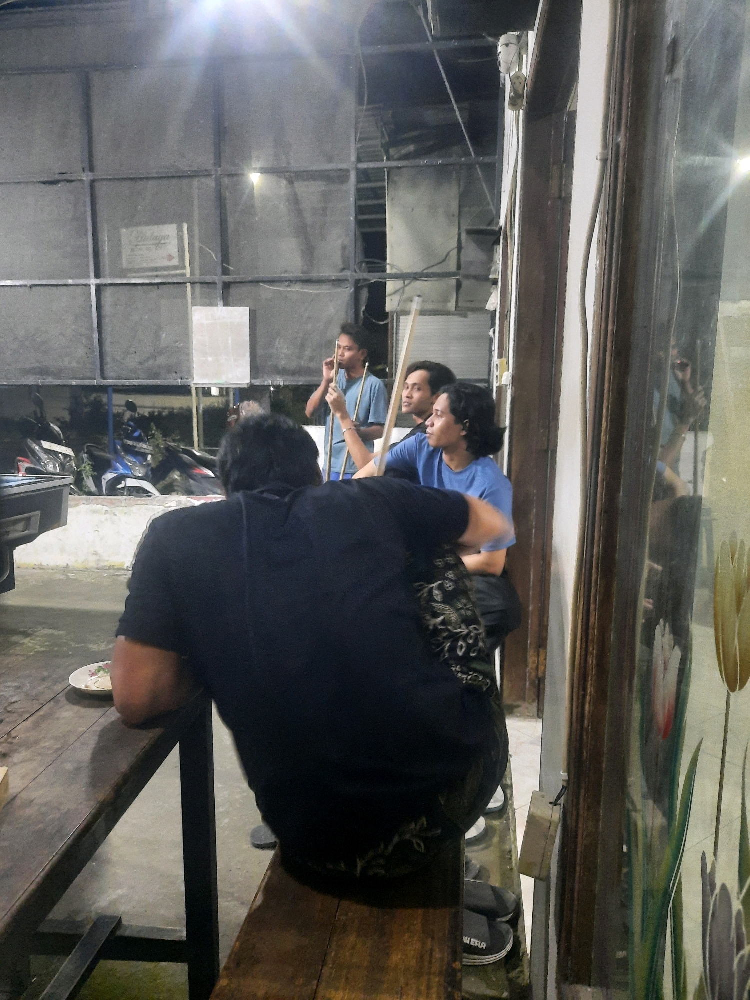 The image depicts an indoor scene in Pucangtelu, Indonesia, possibly a pool hall or a casual gathering spot. In the foreground, a person wearing a black t-shirt and patterned pants is seated at a wooden table, their back to the camera. To their right, two other individuals are visible, one in a blue t-shirt holding a pool cue, and another person beside them. In the background, there are motorcycles parked and a large, gridded structure. A reflective surface on the right side of the image reveals a partial reflection of the individuals and the environment. The lighting suggests an evening or dimly lit setting, with a bright light source visible in the upper portion of the frame. The overall atmosphere appears relaxed and informal.