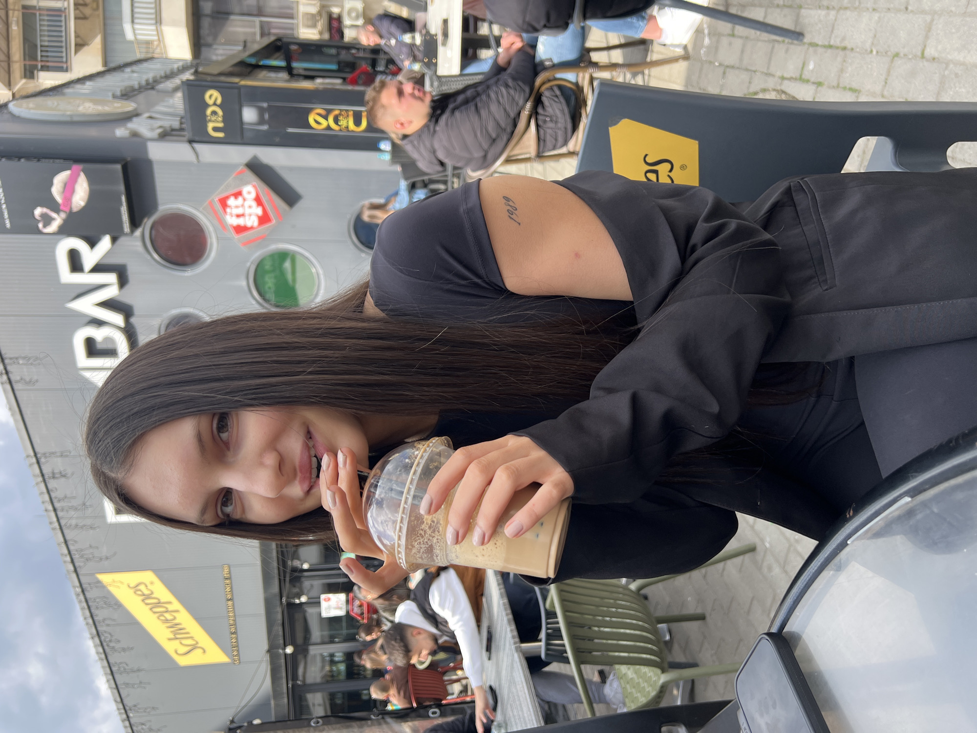 A young adult female with long, dark hair is seated at an outdoor cafe table, actively drinking from a clear plastic cup containing a light brown beverage, likely iced coffee or boba tea, through a straw. She is dressed in a black short-sleeved top and a black blazer, with light-colored nail polish. A tattoo displaying the numerical sequence "1989" is visible on her left upper arm. Her gaze is directed towards the camera, and her lips are slightly parted around the straw. On the glass-topped table to her left, a dark-colored smartphone is present. She occupies a dark grey plastic chair.
The background reveals a bustling outdoor commercial street with additional patrons seated at adjacent tables. The building facade behind the subject prominently displays multiple commercial signs. These include a large yellow "Schweppes GENUINE SUPERIOR SINCE 1783" sign, a white "BAR" sign, a black sign with a pink heart graphic and "CIE", a red and white "fit spo" sign, and a vertical black sign with gold text identifying "ECU cafe". The setting is a paved pedestrian area under a partially cloudy sky. This scene is located on Vitosha Boulevard in Sofia, Bulgaria, characterized by the identifiable "BAR ECU" and "fit spo" establishments.