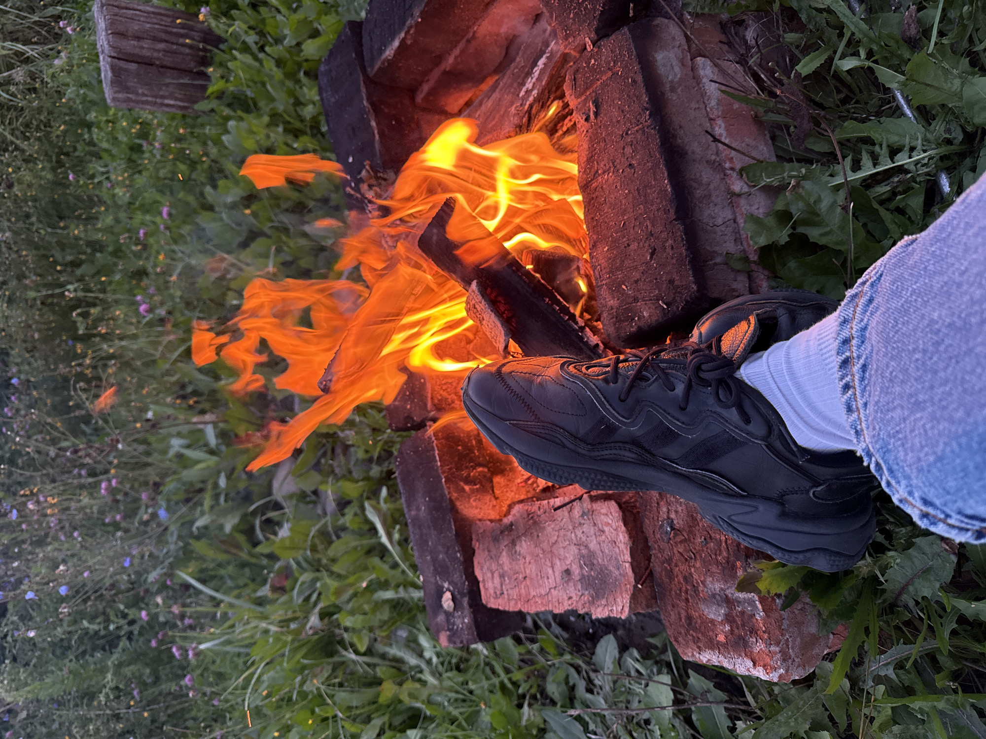 A pair of black athletic shoes and blue jeans are positioned in the foreground, with the right shoe resting on a makeshift fire pit constructed from red bricks. The fire pit contains a bright orange flame, with burning logs and embers visible. The background consists of a natural environment with green grass, weeds, and small wildflowers. A wooden post is visible to the right of the fire. The overall scene suggests an outdoor setting, possibly a campsite or backyard. The location is identified as Imelevo, Russia.