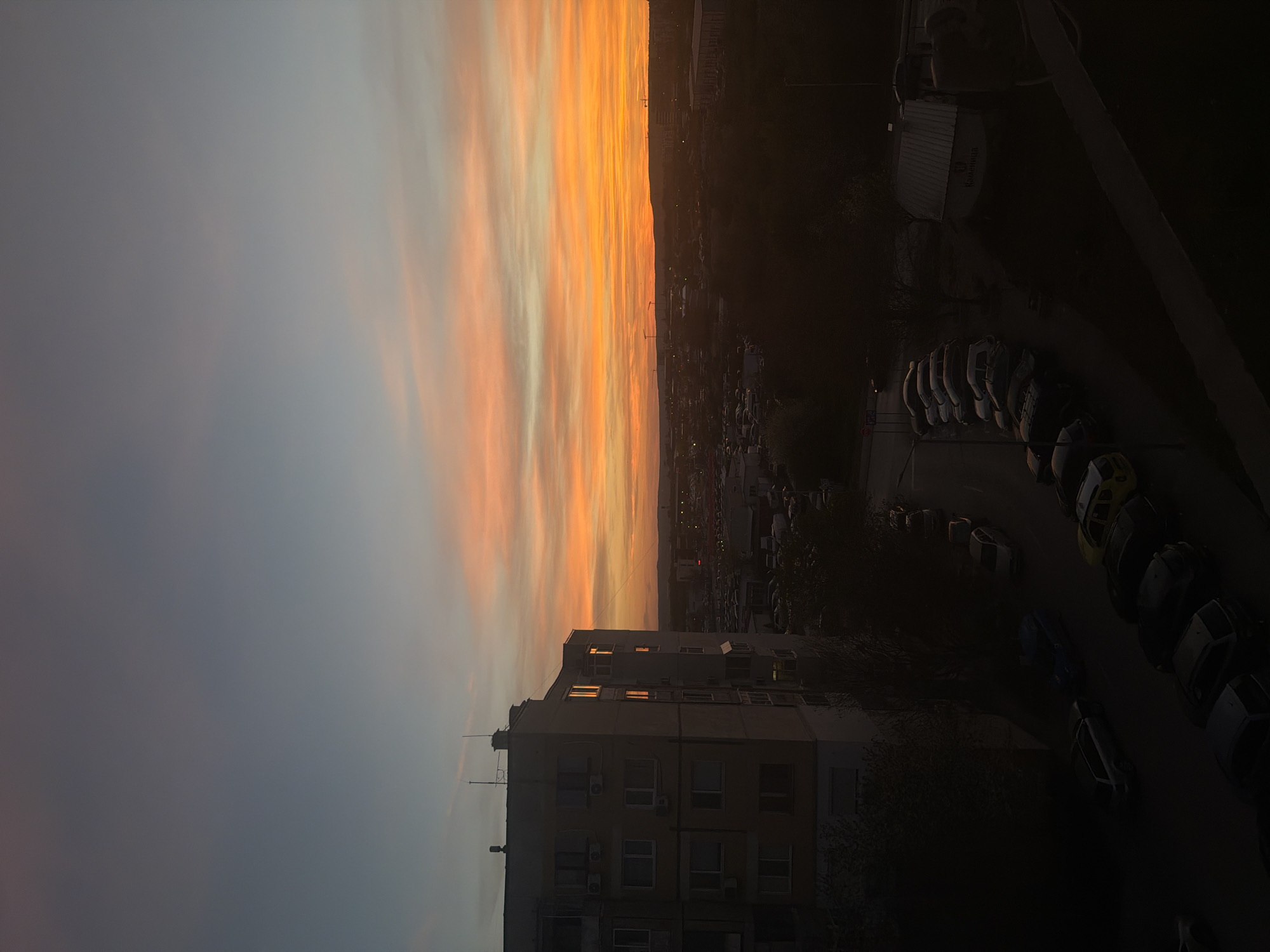 The image depicts an urban landscape at sunset, captured from a elevated vantage point. The sky dominates the upper and left portions of the frame, showcasing a spectacular gradient of colors. Vibrant streaks of orange, yellow, and red clouds, illuminated by the setting sun, transition into softer pinks and blues, and finally to a muted grey-blue on the right side of the sky.
Below the horizon line, the silhouette of a city stretches into the distance. In the immediate foreground, a multi-story, grey apartment building stands prominently, with some of its windows reflecting the warm hues of the sunset. Below the building and stretching into the dark lower right of the image is a street lined with numerous parked cars, mostly visible as dark shapes against the fading light. One white van or truck is discernible, with the brand name "Husqvarna" visible on its side. Sparse, dark foliage of trees can be seen between the buildings and cars. The overall scene suggests the transition from day to evening in a residential or mixed-use urban area.