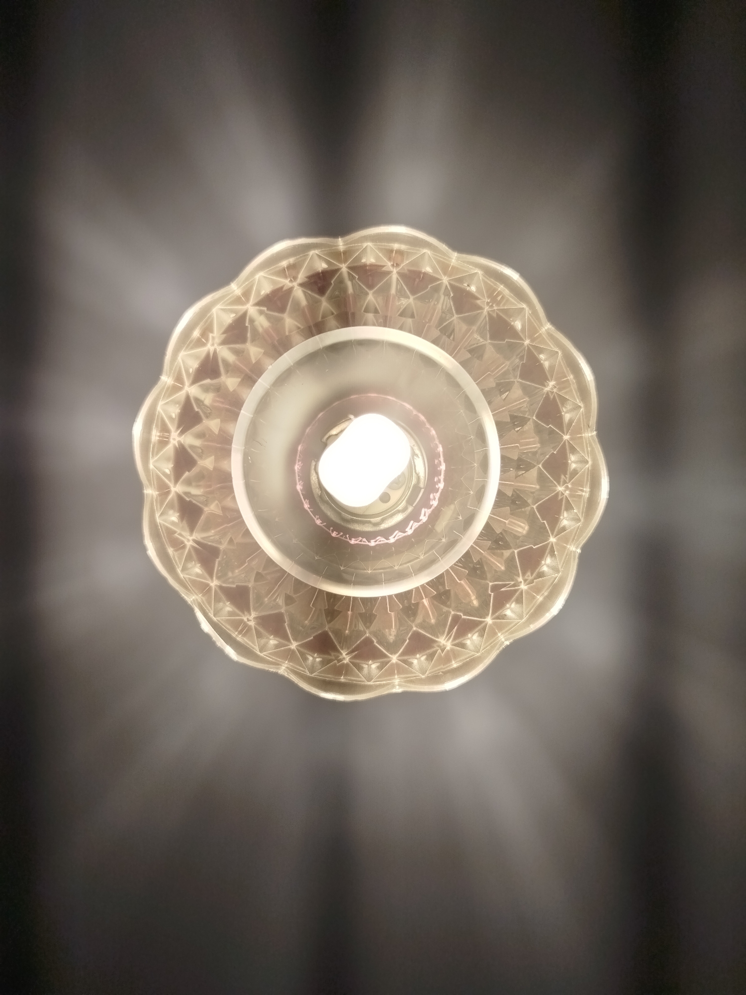 A ceiling lamp with a decorative glass shade is illuminated, casting rays of light onto a dark, textured ceiling. The lamp has a scalloped edge and a geometric pattern on its shade. A bright LED bulb is visible at the center. The scene is devoid of people and any specific context relating to Votkinsk, Russia.