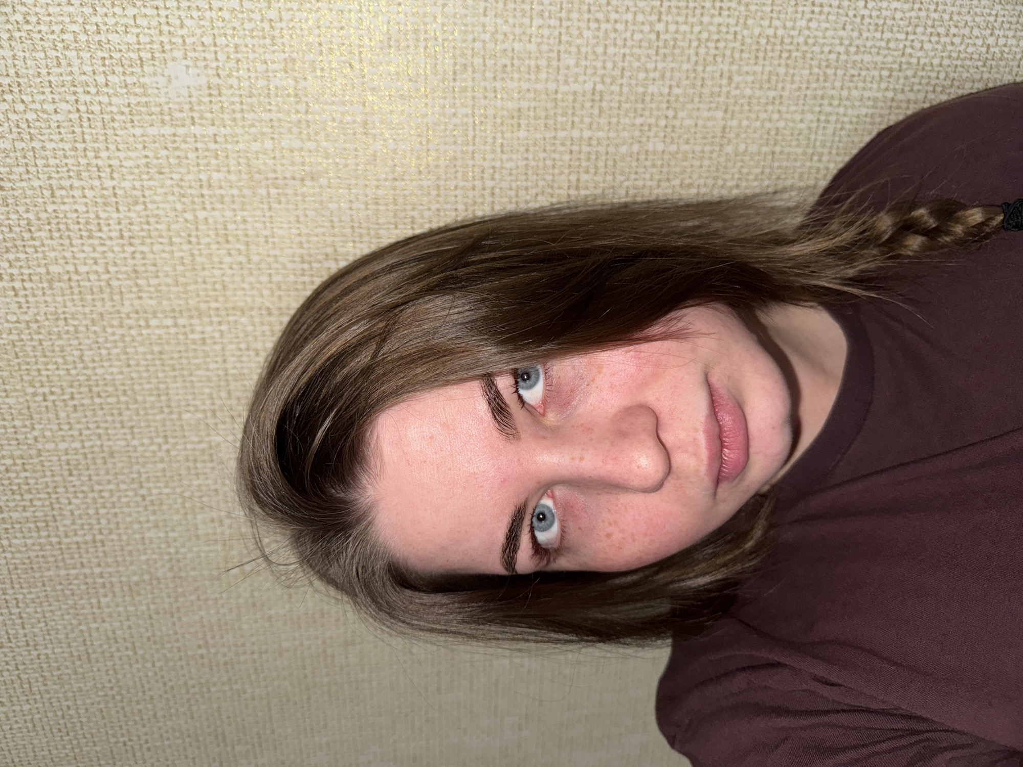 A close-up, angled shot of a person with light skin, freckles, and light brown hair, with a small braid visible on the right side. The person has blue eyes and is wearing a maroon t-shirt. The background is a textured beige wall. There is no information to suggest this photo was taken in Chaikovsky, Russia.
