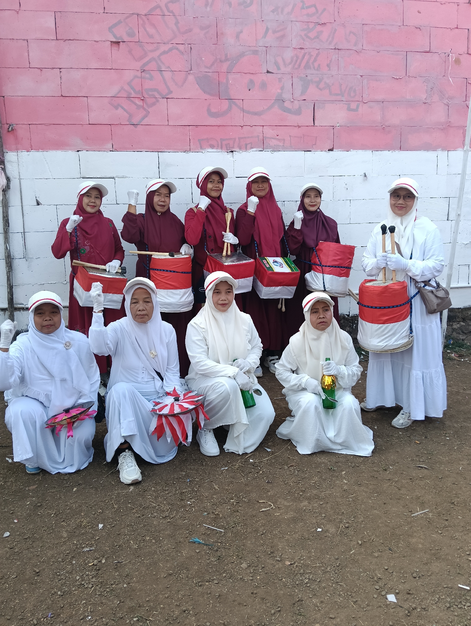 In Tasikmalaya, Indonesia, a group of ten individuals, appearing to be adult women, are posing for a photograph in front of a partially painted brick wall. The wall is adorned with what seems to be spray-painted graffiti in black and pink. The lower portion of the wall is white, creating a striped effect with the pink upper section.
The individuals are dressed in either maroon or white religious attire, consisting of long dresses and headscarves, some complemented by white caps. Seven of them are holding drumsticks and appear to be part of a drumming group. Several drums, wrapped in red and white fabric, are visible, some hanging from the participants' shoulders. Three individuals in the front row are seated, with two of them holding small, red and white striped objects that resemble pinwheels. One seated individual in white is holding a green bottle. The individuals are wearing white gloves and some are raising their fists, suggesting a celebratory or determined stance. The ground beneath them is a dirt and gravel surface. The setting appears to be outdoors, possibly in a semi-urban or rural area, given the rough ground and unfinished wall.