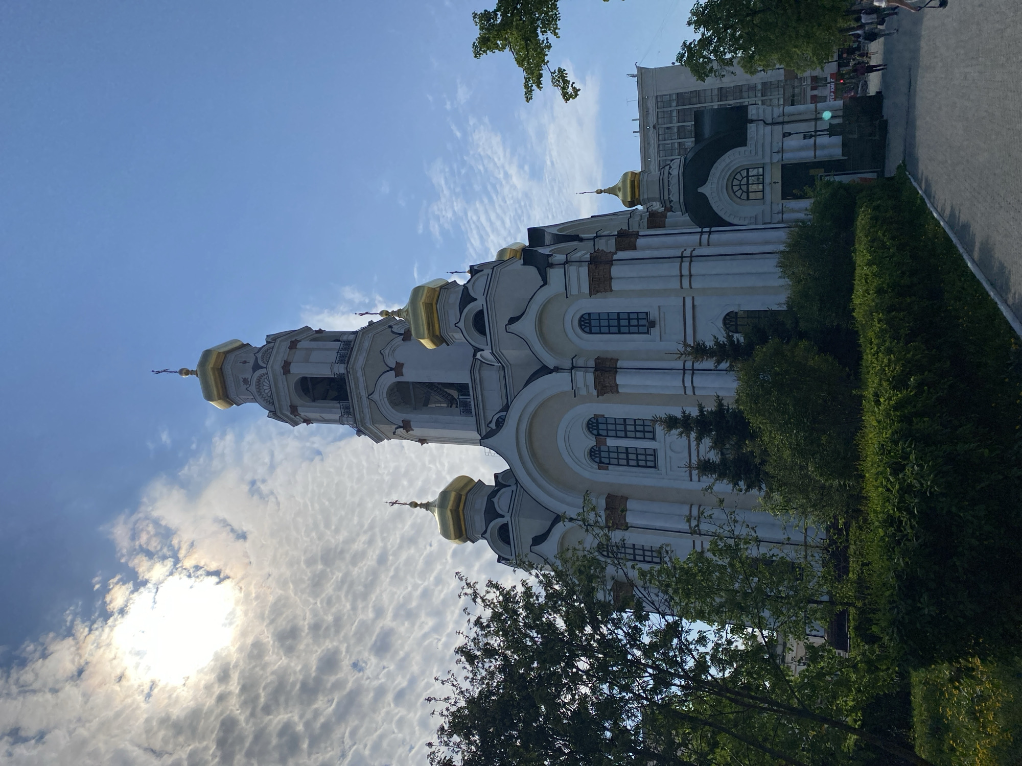 The image depicts the Cathedral of the Ascension in Yekaterinburg, Russia. The cathedral is white with gold domes, standing tall against a partly cloudy blue sky. The sun shines through the clouds. Trees and greenery are in the foreground, partially obscuring the base of the building. A paved walkway and a building with windows are visible to the right of the cathedral. Some people are seen walking along the walkway.
