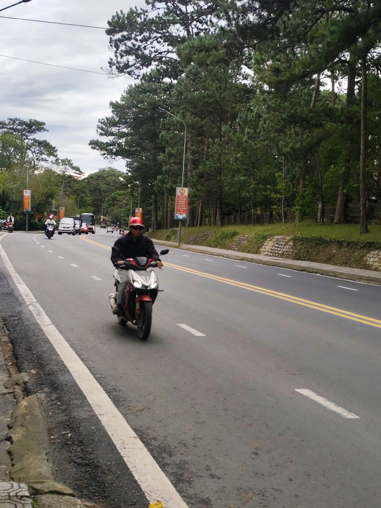 A man wearing a red helmet and a dark jacket rides a red and white motorcycle on a paved road in Da Lat, Vietnam. The road has a white line marking the shoulder and two yellow lines marking the lanes. Trees line the right side of the road, with a retaining wall made of stone blocks visible below the trees. In the background, several other vehicles, including cars and buses, are traveling on the road. Banners with Vietnamese text are posted on poles along the roadside. The sky is overcast.