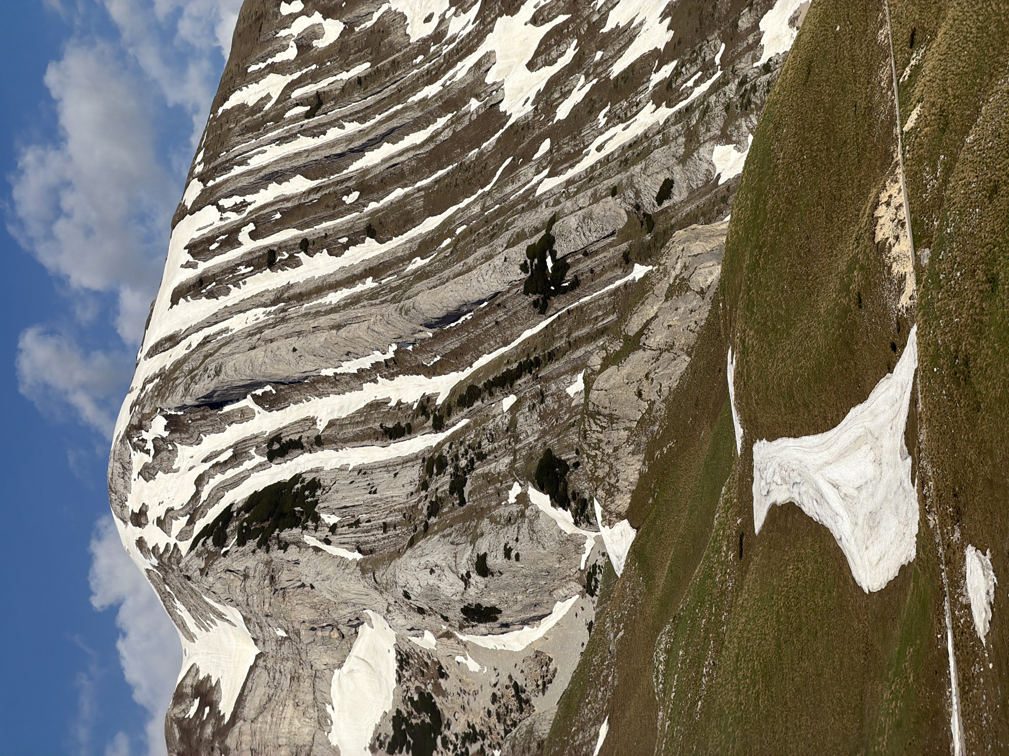 An aerial view depicts a mountain landscape in Боричје, Montenegro. A prominent, layered rock face dominates the left side of the image, its horizontal strata accented by patches of snow. The upper portion of the face is juxtaposed against a blue sky with scattered clouds. The right side of the image features a grassy slope interspersed with snow patches, creating a contrast to the bare rock face.