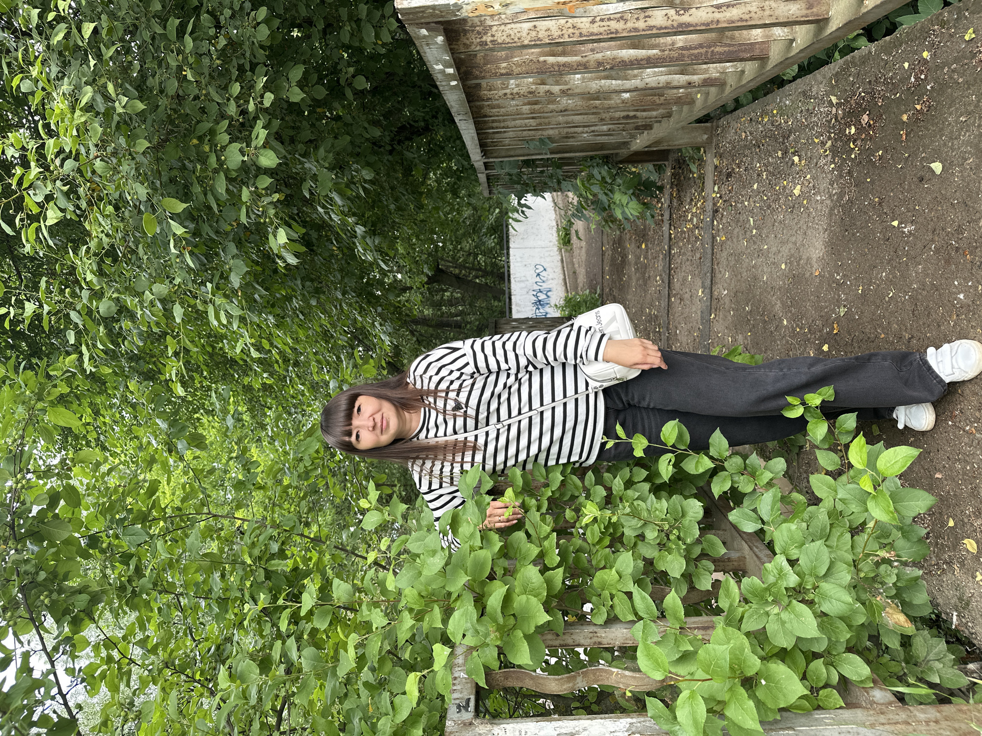 A young woman with dark hair and bangs stands on a weathered wooden bridge in Chaikovsky, Russia. She is wearing a black and white striped long-sleeved shirt, dark pants, and white sneakers. A small white purse is slung across her body. She is leaning on a rusted metal fence, partially obscured by lush green foliage. The bridge is made of wood and concrete, with fallen leaves scattered on its surface. In the background, a white wall with graffiti is visible, and dense green trees surround the area, suggesting a natural or park-like setting within the city. The lighting is overcast, indicating a cloudy day.