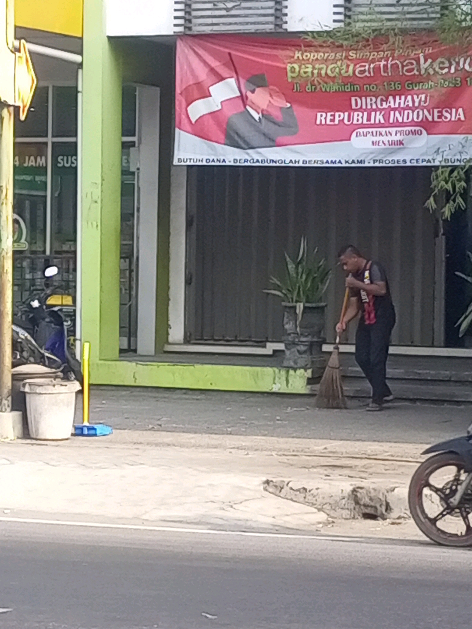 A street scene in Gurah, Indonesia, depicts a man sweeping the sidewalk in front of a building. The building has a green-painted facade and a large red banner that reads "Koperasi Simpan Pinjam panquarthaken" and "DIRGAHAYU REPUBLIK INDONESIA" along with an address. To the left of the building, a yellow directional arrow sign is visible. A motorcycle is parked on the left side of the frame, next to a large trash bin. The man, wearing a black shirt with red accents and black pants, is actively sweeping with a broom. He is positioned on the sidewalk, with a potted plant behind him. The street in the foreground shows asphalt with a white line.