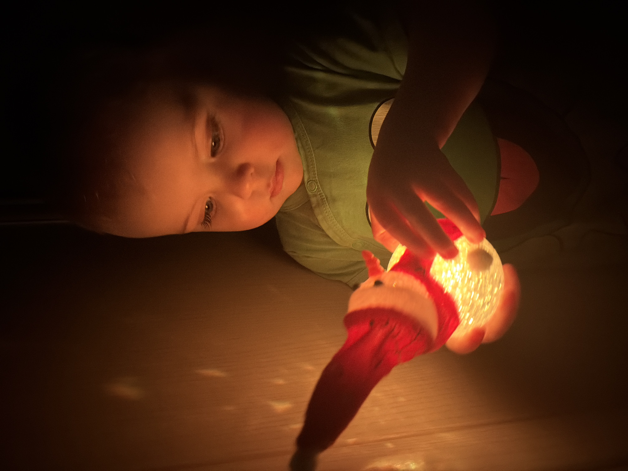 A young child wearing a green shirt with a round design is lying down and holding a lit Santa Claus-themed light. The child's face is illuminated by the light, which also casts a glow on the surrounding surface. The scene appears to be indoors and dimly lit. There is no information to establish the location of the image within Votkinsk, Russia, or to confirm that it is located there at all.