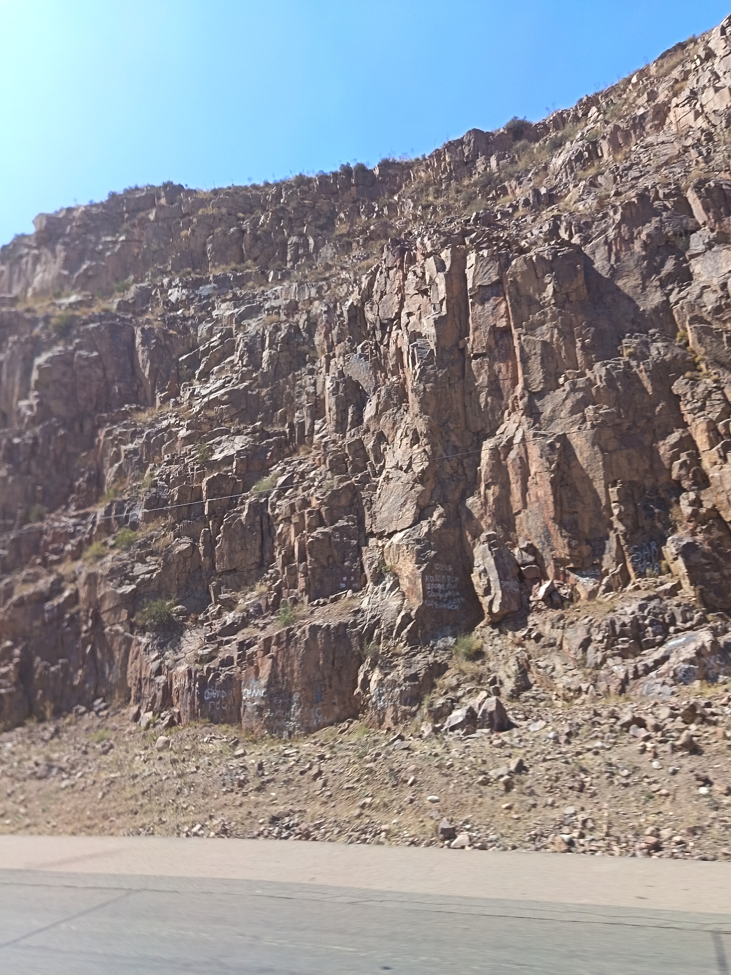 The image shows a steep, rocky cliff face under a clear blue sky. The cliff is composed of large, angular rocks in shades of brown and grey, with some patches of green vegetation growing on ledges and at the base. There is evidence of graffiti scribbled on several sections of the rock face. The base of the cliff meets a dry, rocky slope, which in turn leads to a paved surface in the foreground, suggesting it is a roadside view. The scene appears to be a natural landscape feature near a road. The image does not show any people or specific landmarks that would definitively place it in Beshkul, Uzbekistan.