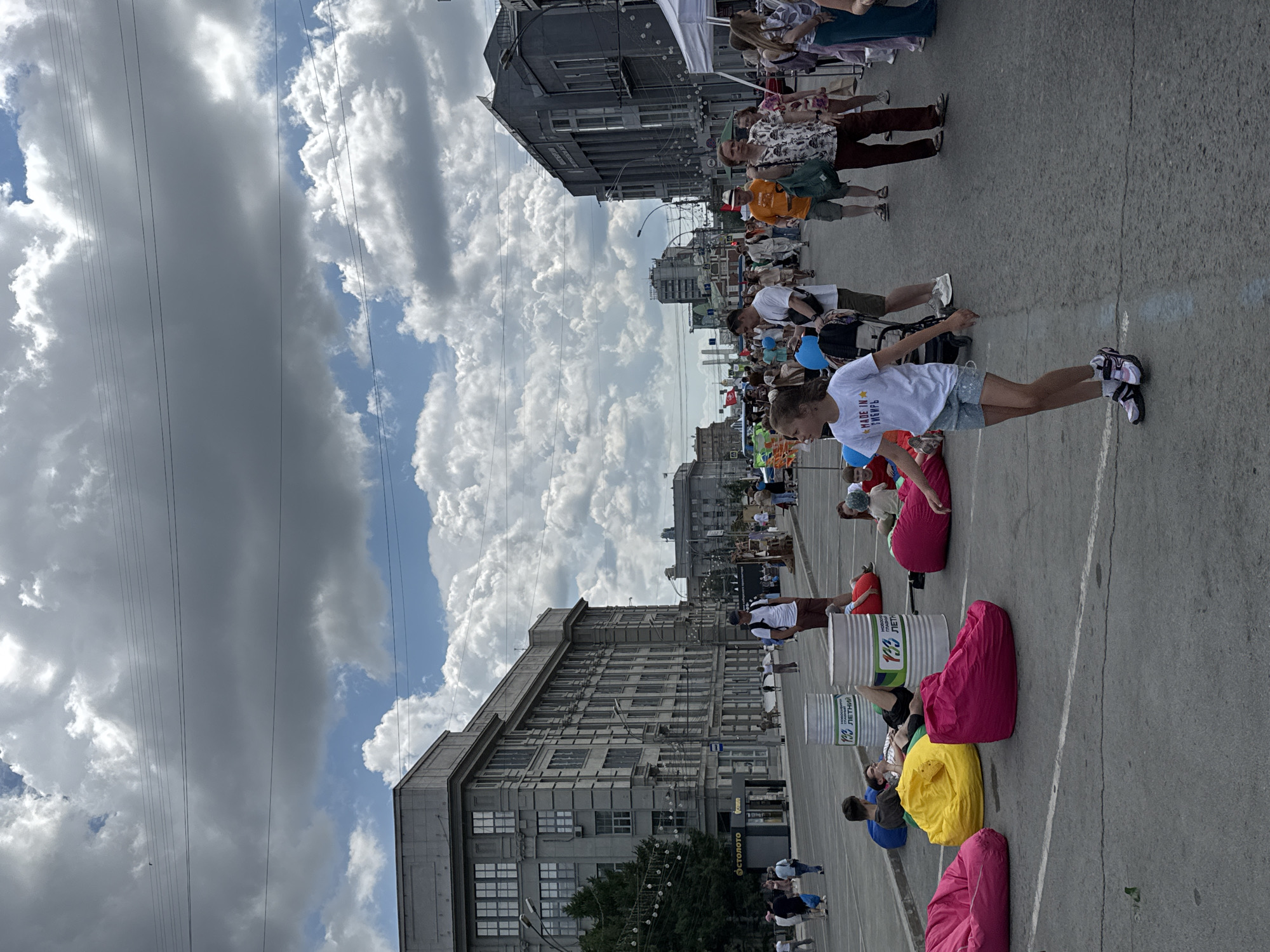 This image depicts a street festival or event in Novosibirsk, Russia, likely celebrating the city's 130th anniversary, as indicated by text on cylindrical objects in the foreground. A large crowd of people are present on a paved street, with buildings lining both sides.
In the foreground, several large, brightly colored beanbag chairs are scattered across the pavement. Some people are seated or lying on these beanbags, relaxing. Two large cylindrical objects, resembling industrial barrels, are also in the foreground, with text in Russian that includes "130 ЛЕТНИЙ" (130th anniversary) and "НОВОСИБИРСКУ" (to Novosibirsk).
A young girl in a white t-shirt with the text "MADE IN SIBIR" and blue shorts is walking in the middle of the street, in the right half of the image. She is wearing sneakers and appears to be dancing or moving playfully.
Further into the street, a diverse crowd of people are walking, standing, and interacting. Some individuals are dressed in casual summer clothing. There are tents set up on the right side of the street, suggesting vendor stalls or event booths.
The buildings in the background are a mix of architectural styles, with a large, imposing building on the left and a narrower, more modern-looking building on the right. The sky is partly cloudy, with large, dramatic clouds dominating the upper portion of the image. Electrical wires are visible crisscrossing the sky, typical of an urban environment.
The overall scene suggests a lively and festive public gathering in an urban setting, with people enjoying an outdoor event.