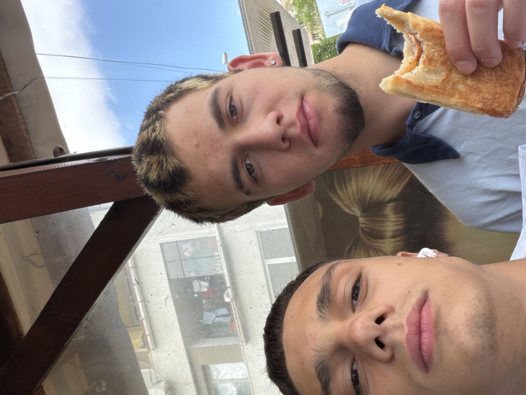 Two men are in the frame. One man is holding a partially eaten pastry. The other man has a wireless earbud in his ear. The background shows buildings, potentially in Varna, Bulgaria.